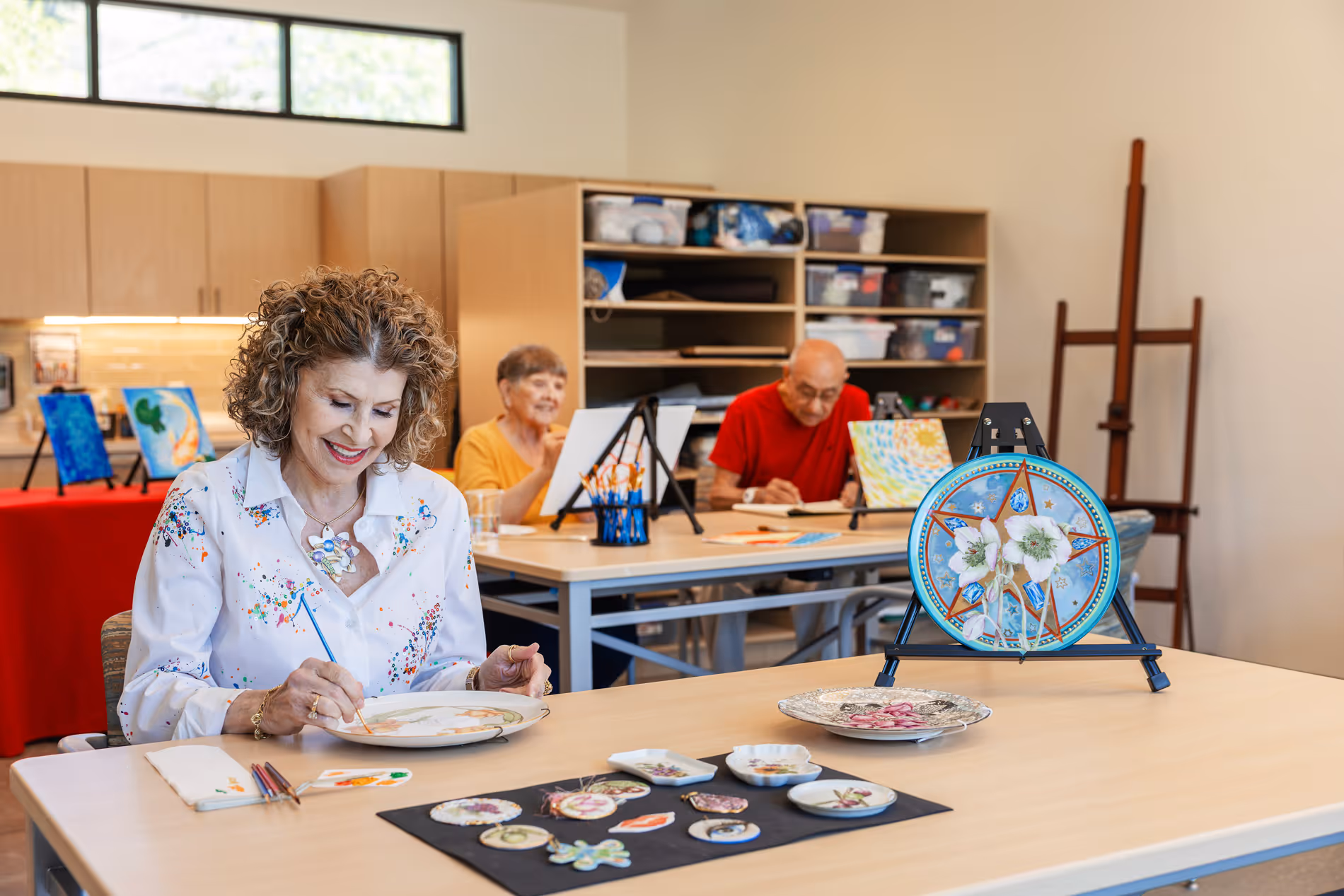 Three seniors painting at tables in an art studio with floral plate designs in the foreground.
