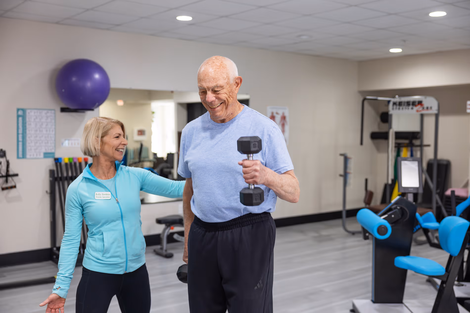 Smiling elderly man lifting dumbbells in a gym while female fitness trainer supports him.