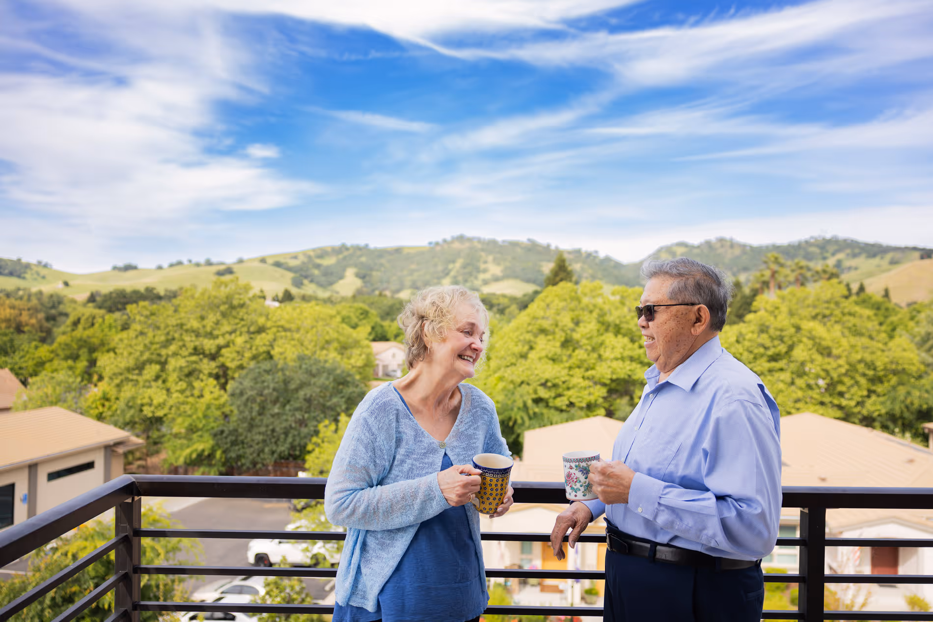 Older man and woman smiling while holding mugs and talking on a balcony with a green hill background.