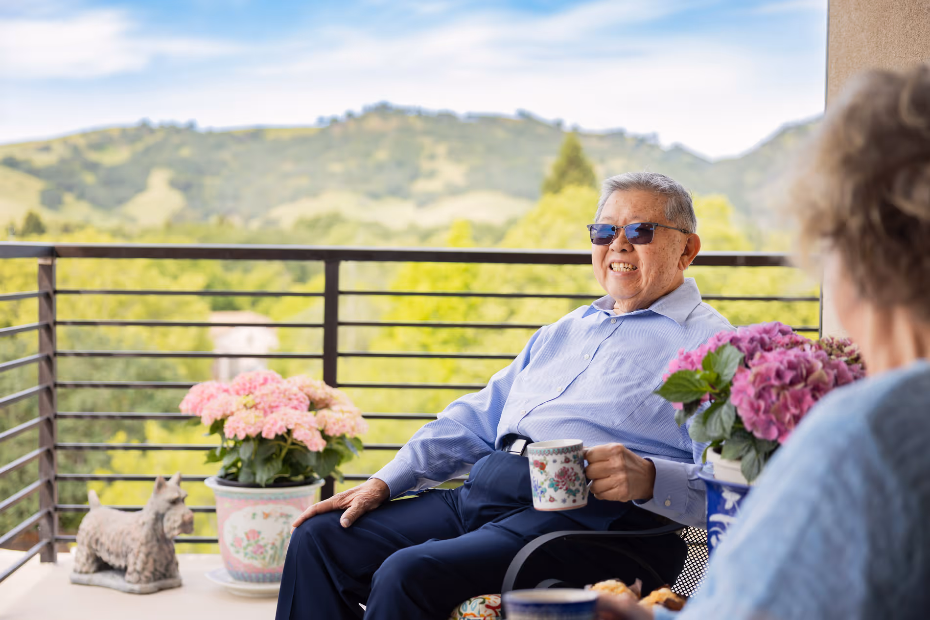 Elderly man smiling with sunglasses holding a floral mug, sitting on a balcony with plants and mountains behind.