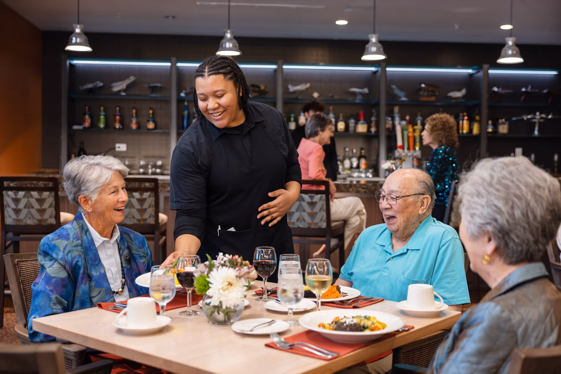 Smiling waitress serving food to three elderly people dining together in a restaurant setting.