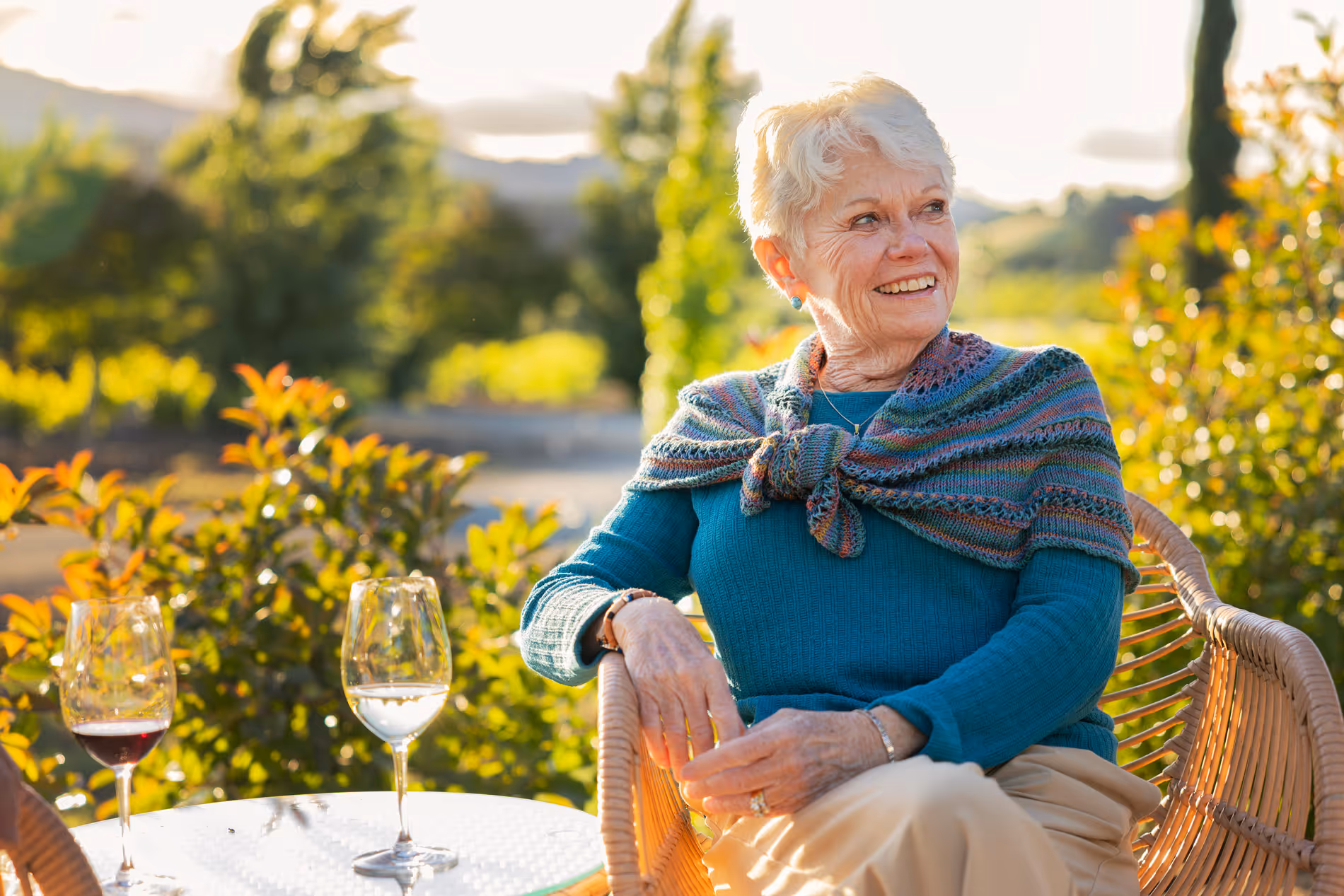 Senior woman in teal sweater sitting outdoors at a table with two wine glasses, smiling.