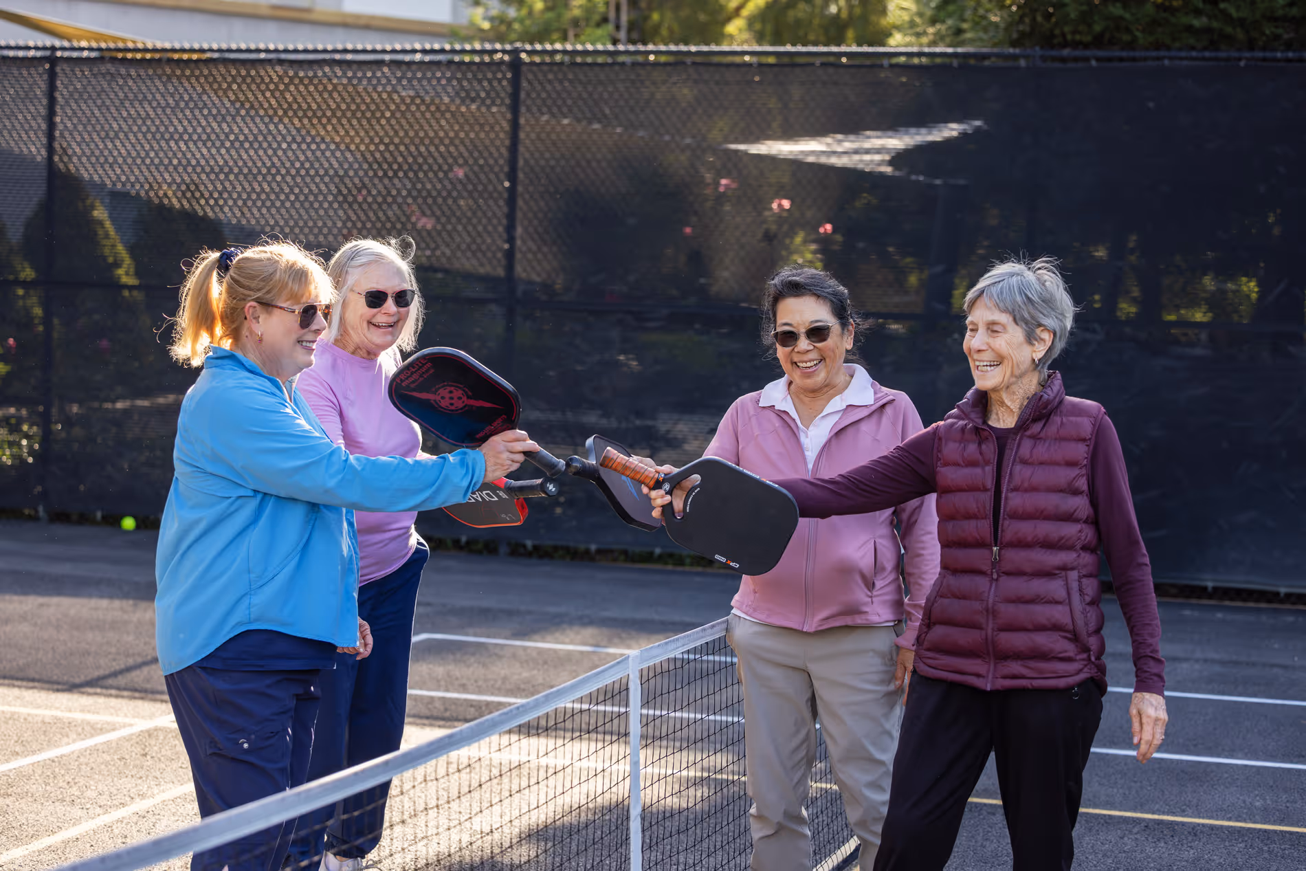 Four smiling older women holding pickleball paddles together over the net on an outdoor court.