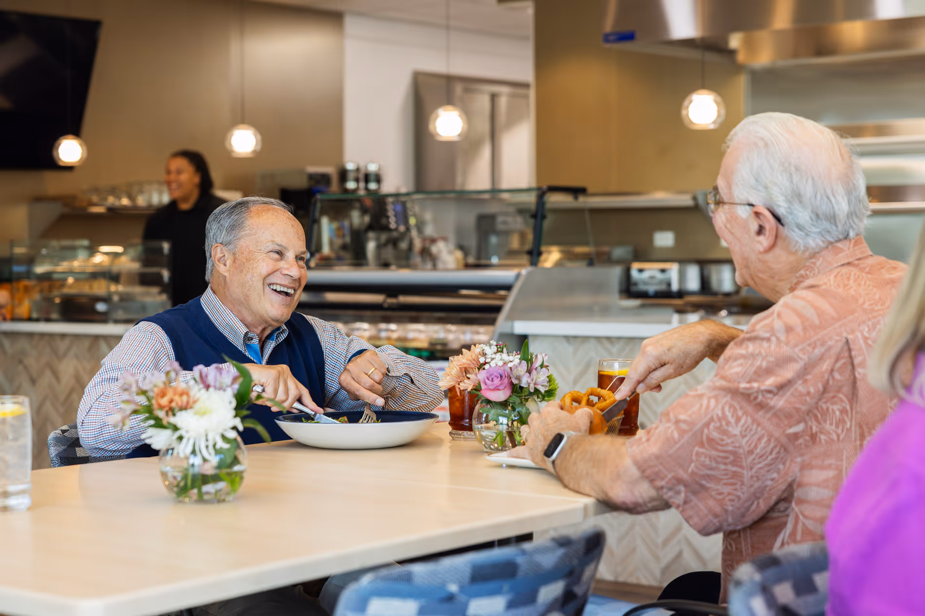 Two older men smiling and eating at a restaurant table with drinks and flower vases.