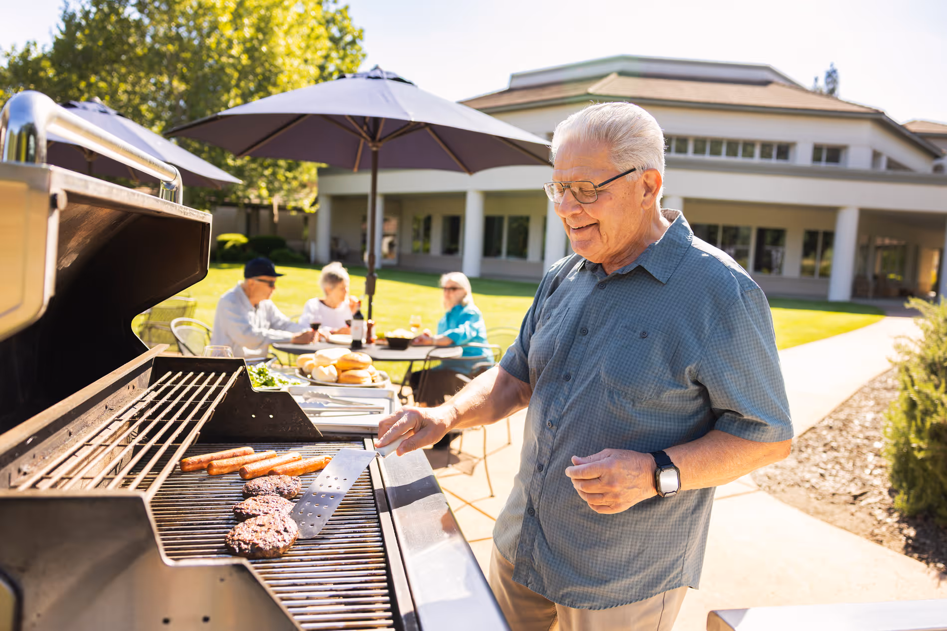 Elderly man grilling burgers and hot dogs outdoors while people sit at a table in background.