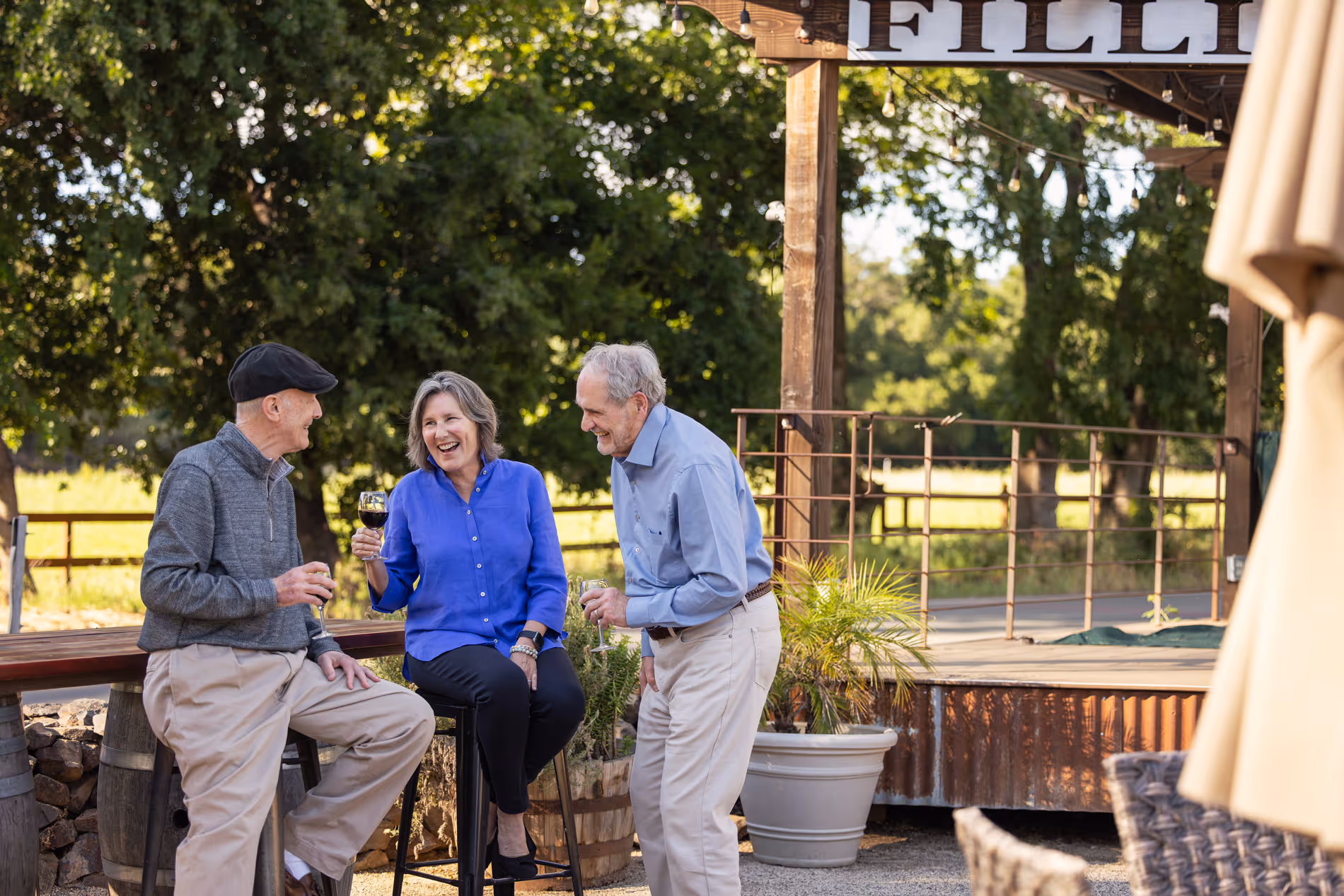 Three older adults outdoors enjoying wine and laughing near rustic wooden seating and greenery.