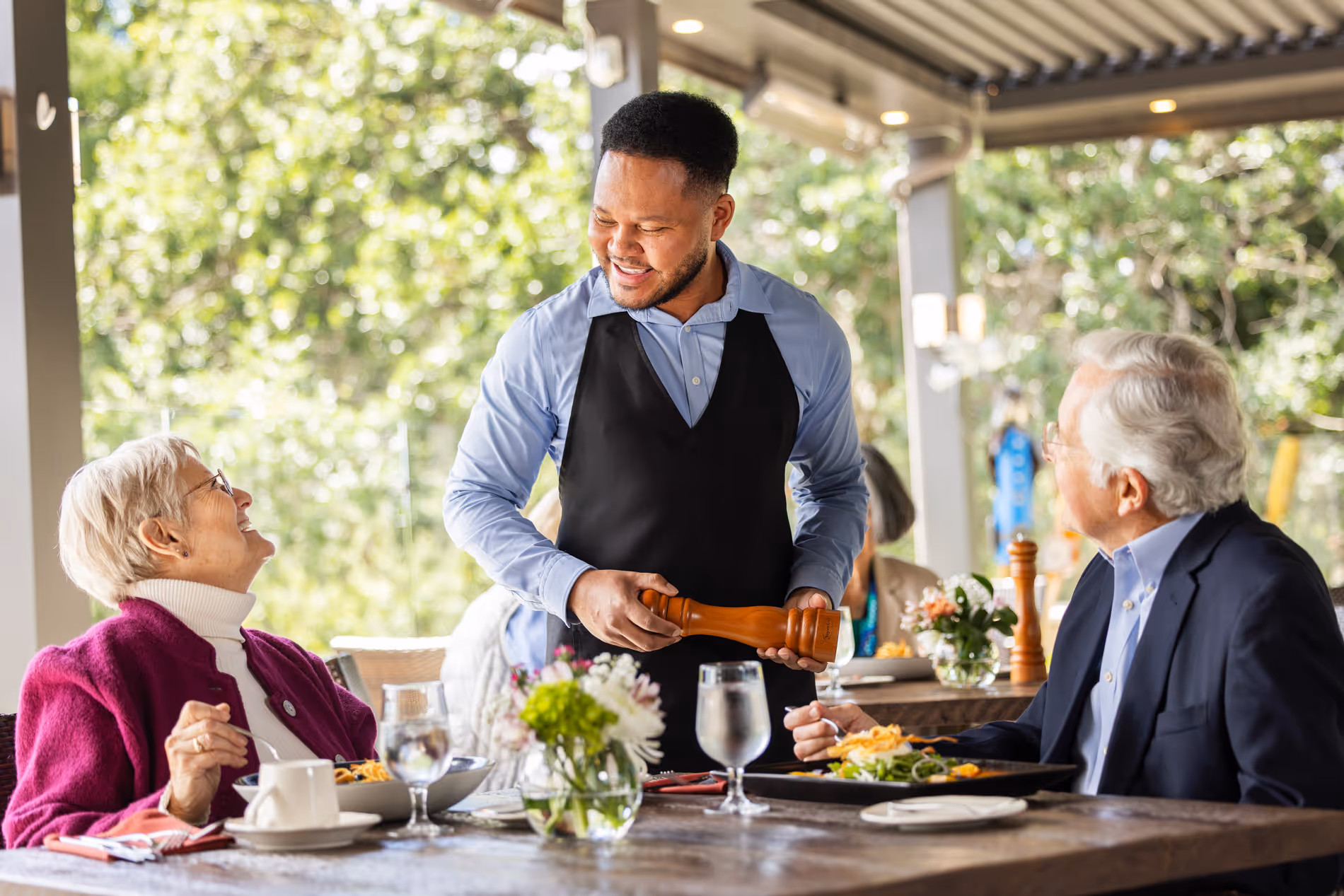 Waiter with pepper grinder serving elderly couple dining outdoors at a restaurant table.