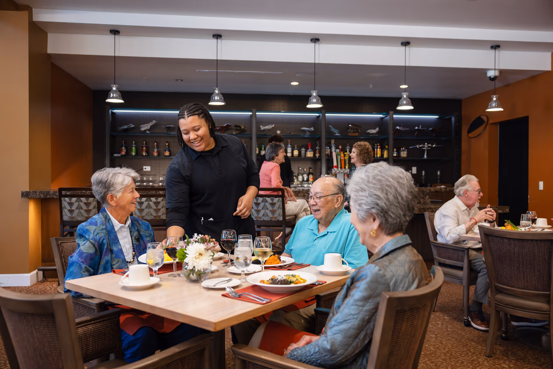Waitress serving food to smiling elderly people seated at a dining table in a cozy restaurant.