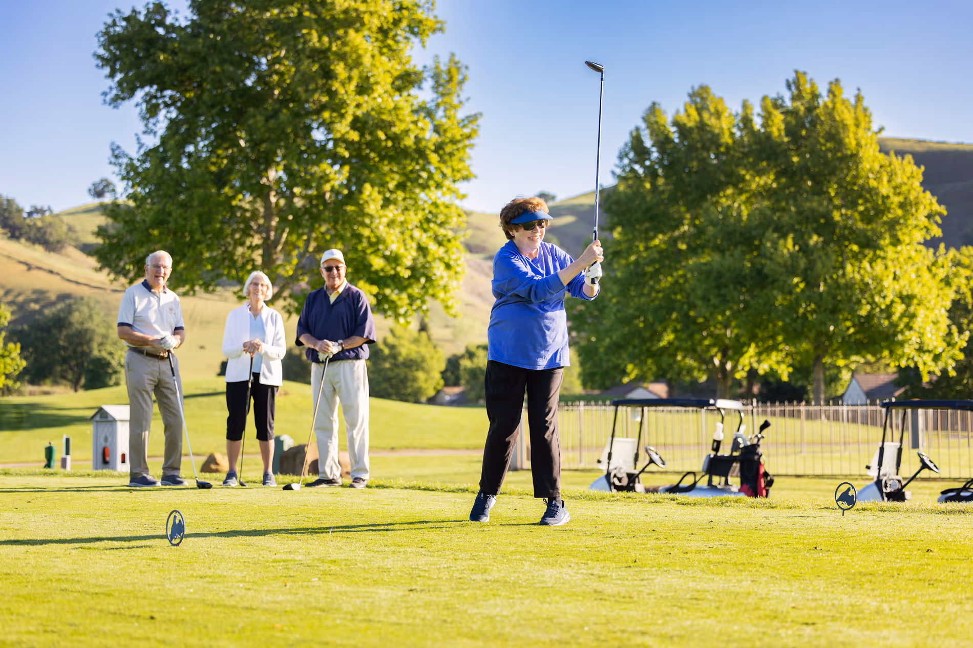 Older woman swinging golf club on course with three seniors watching and golf carts nearby on sunny day.