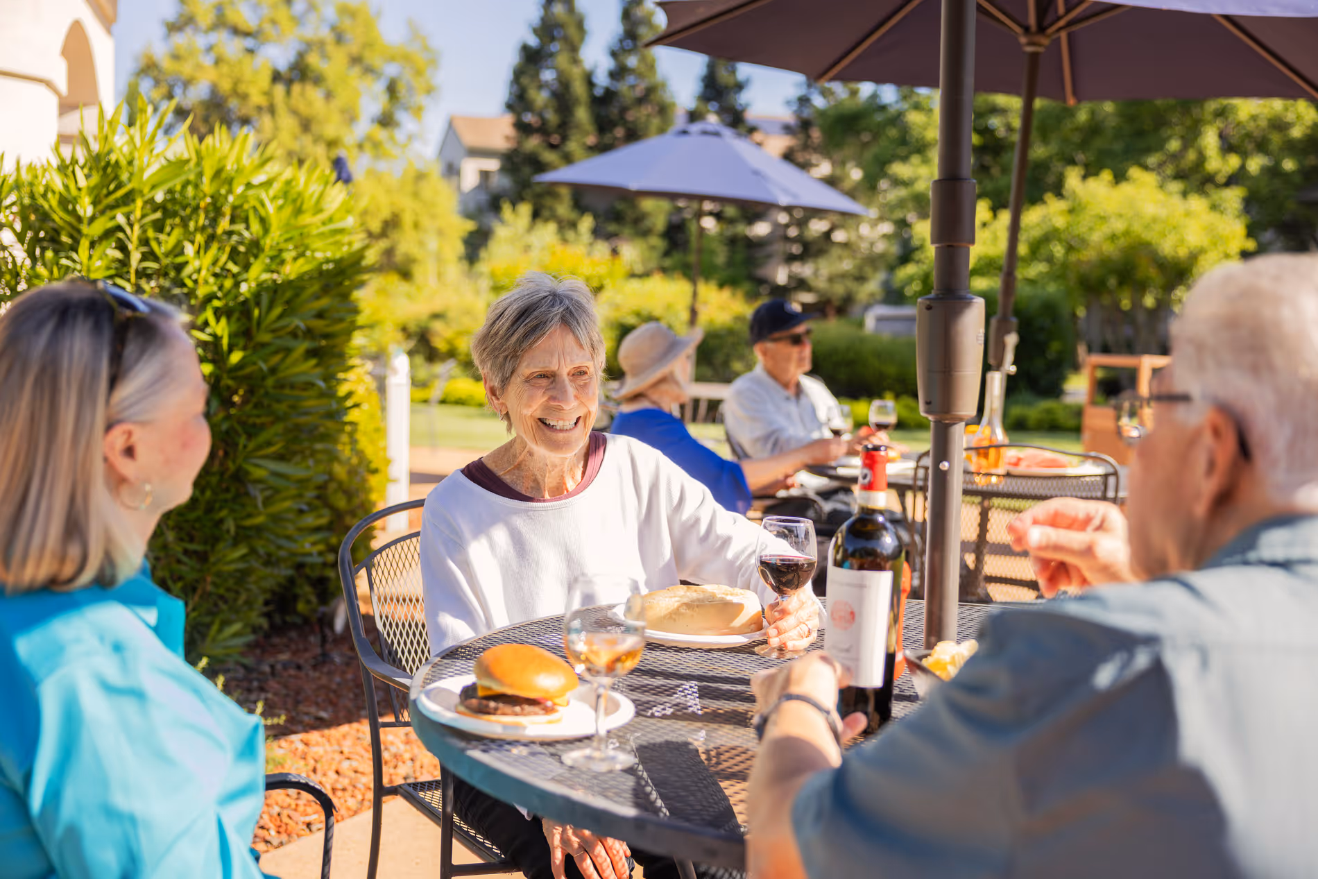 Seniors enjoying outdoor meal with wine and sandwiches under patio umbrellas on a sunny day.