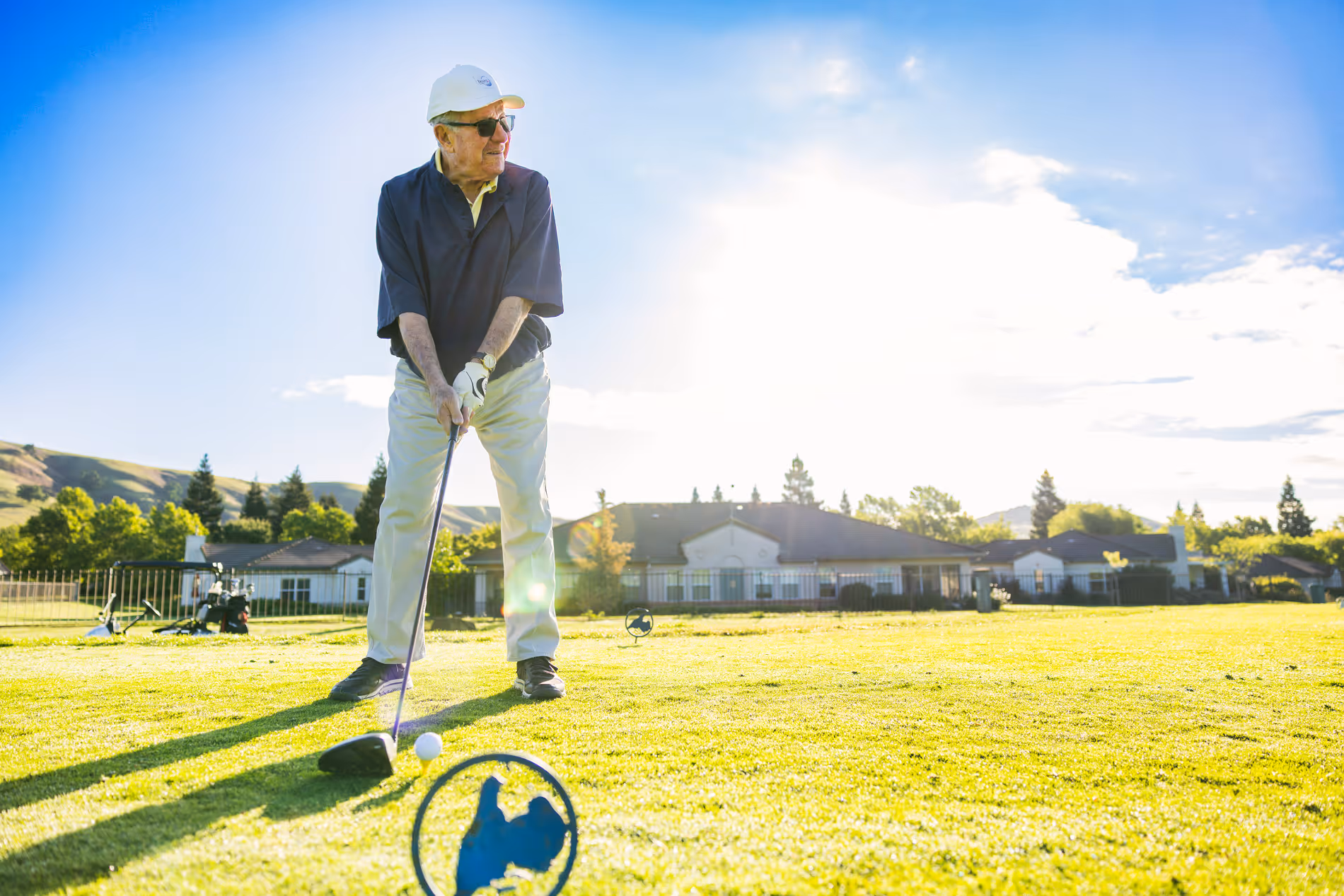 Elderly man in sunglasses preparing to hit a golf ball on a sunny golf course near houses.
