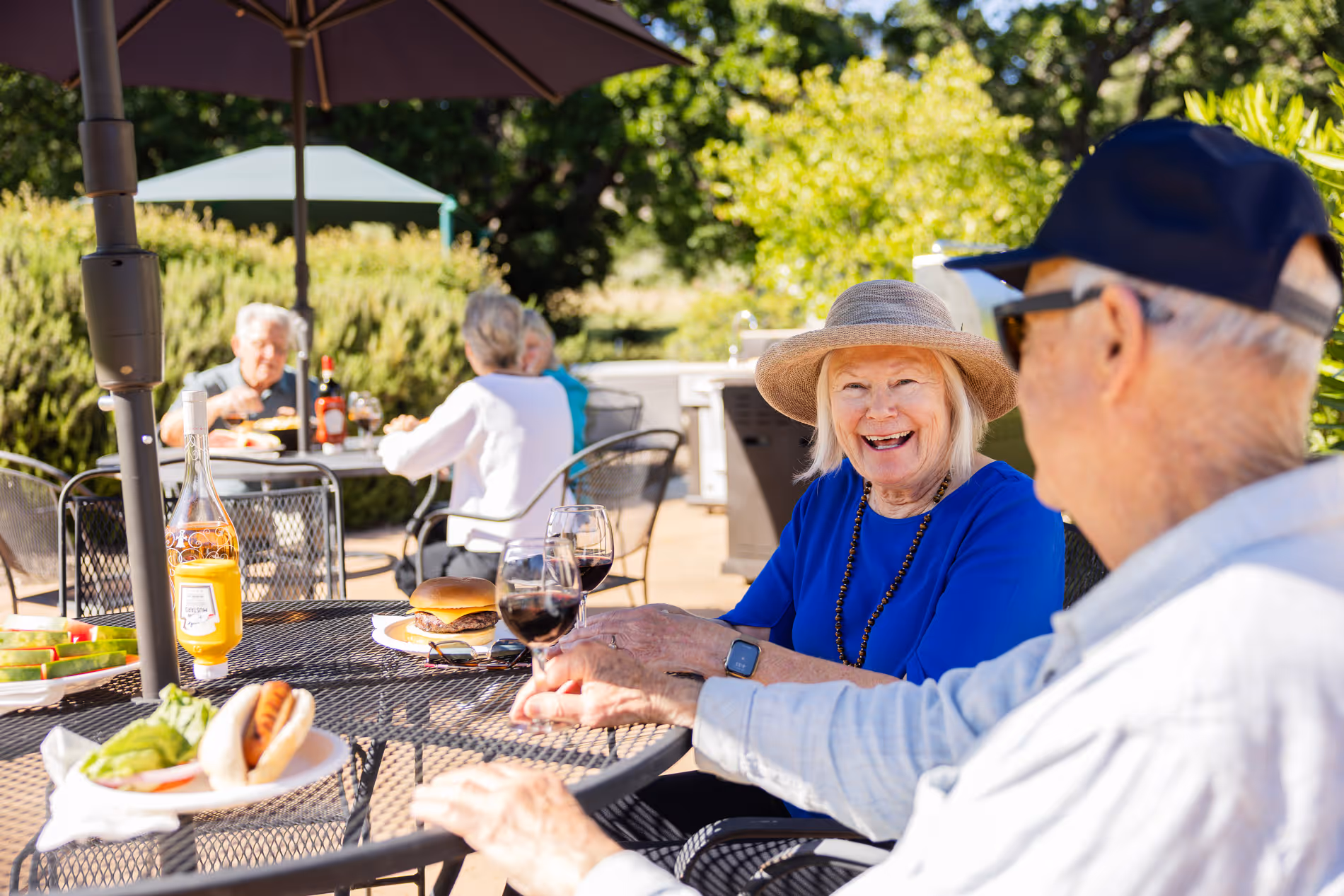 Elderly woman in a hat smiles holding a glass of red wine while sitting outside at a table with food.