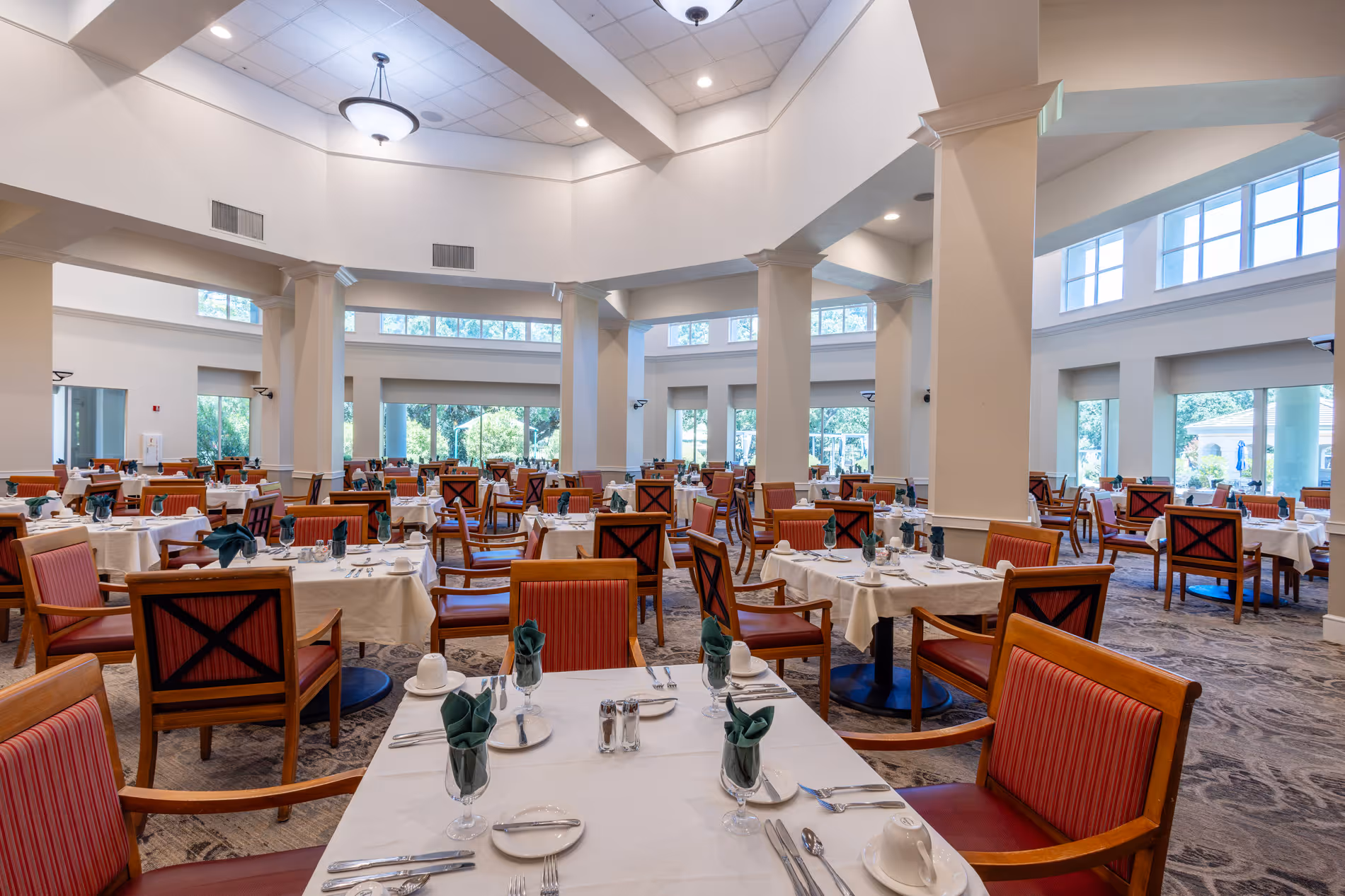 Spacious dining room with multiple set tables, wooden chairs, large windows, and bright lighting.