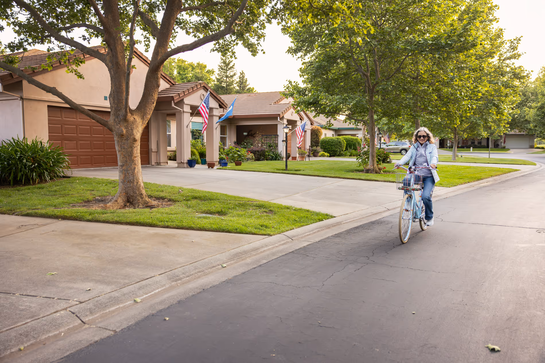 Older woman wearing sunglasses rides a blue bike along a tree-lined suburban street with beige houses.