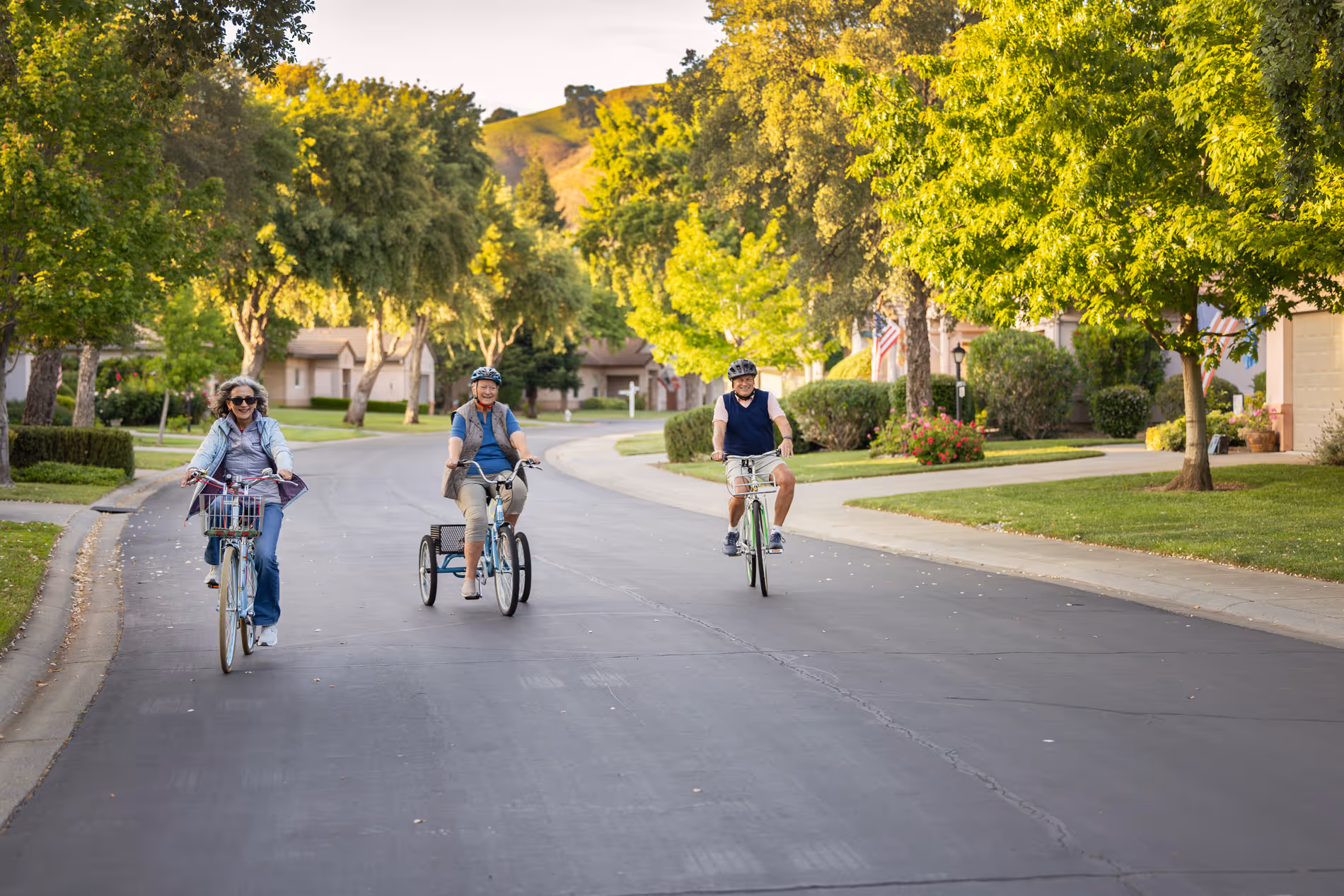 Three seniors riding bikes on a tree-lined suburban street on a sunny day.