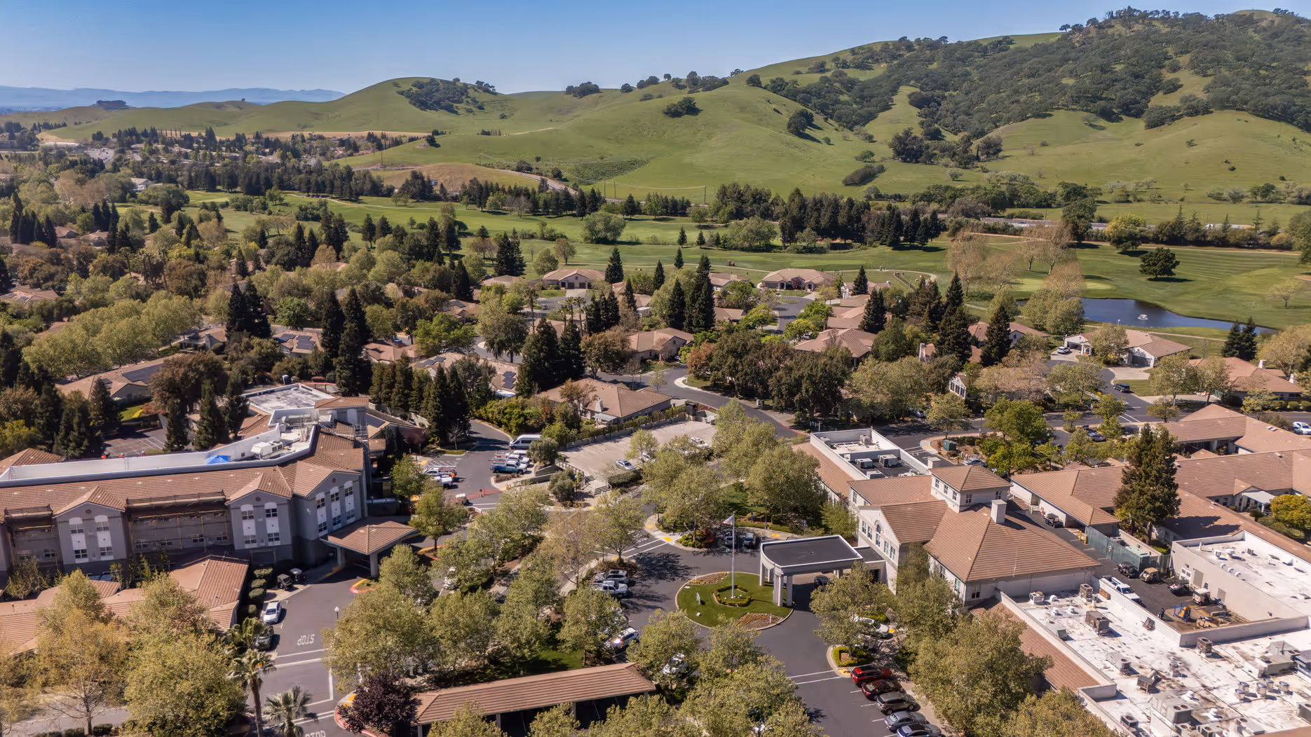 Aerial view of suburban buildings, green trees, parking lots, and rolling hills under a clear sky.