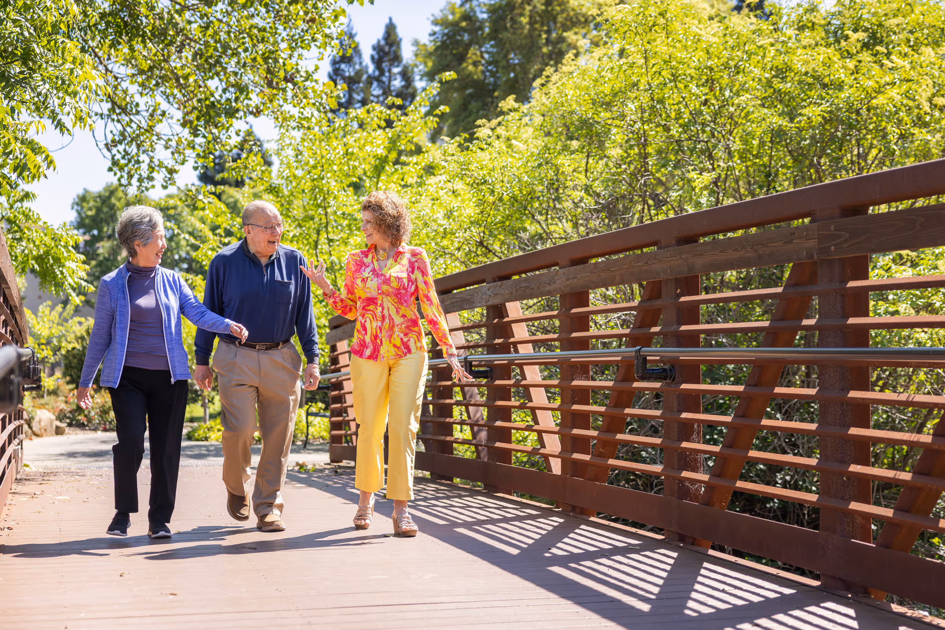 Three adults walking and talking on a sunny wooden bridge surrounded by green trees.