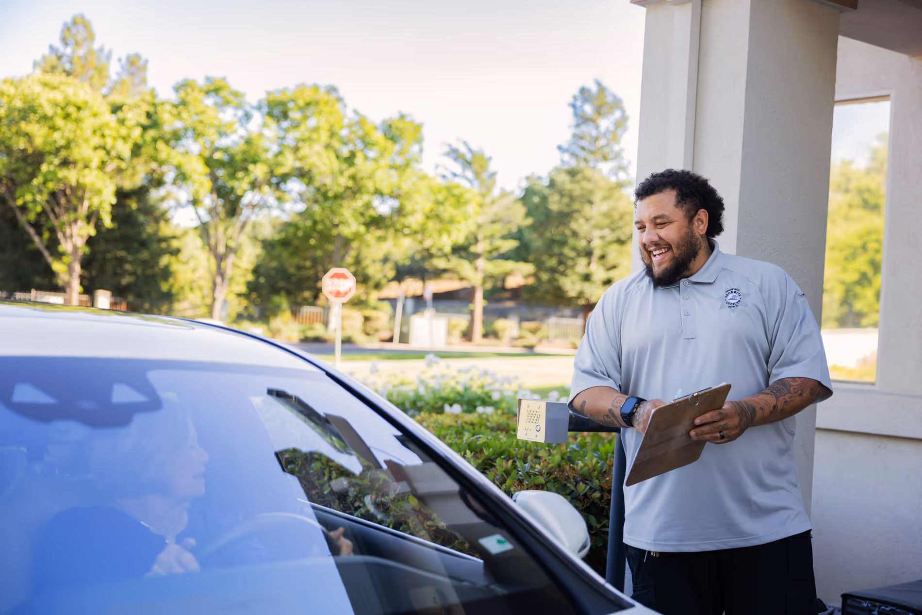 Smiling security guard with clipboard talking to a driver in a white car at a checkpoint.