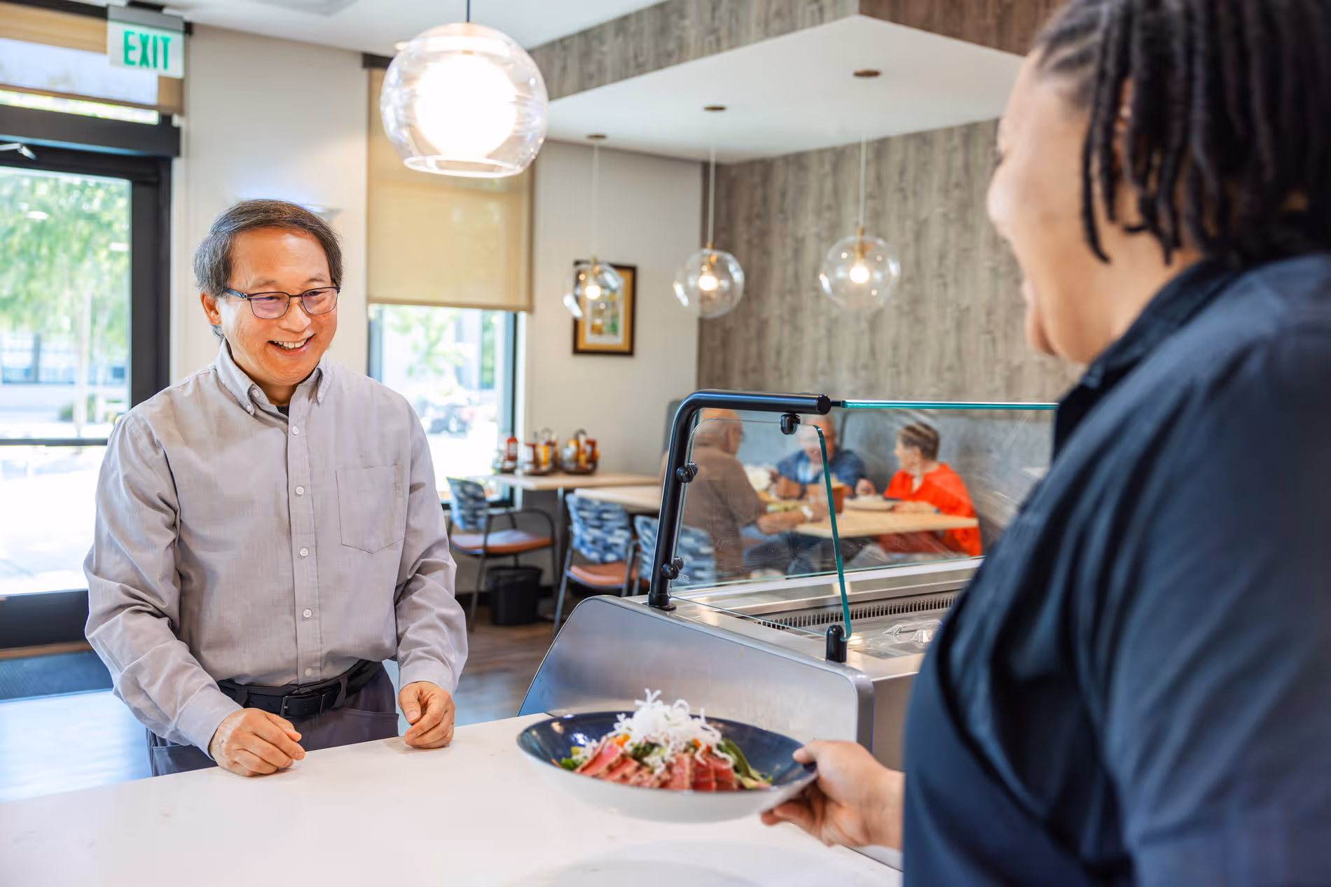 A man smiles as a woman serves him a plate of food at a restaurant counter.