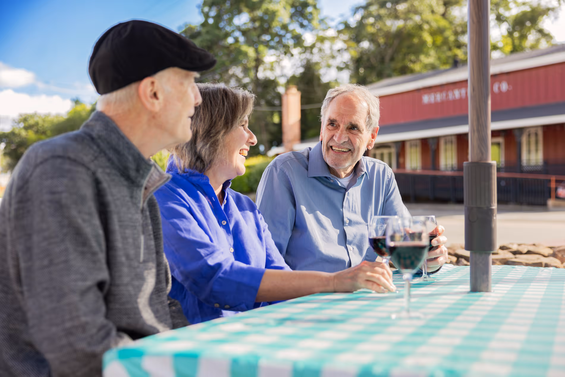 Three people sit at an outdoor table, smiling and holding wine glasses.