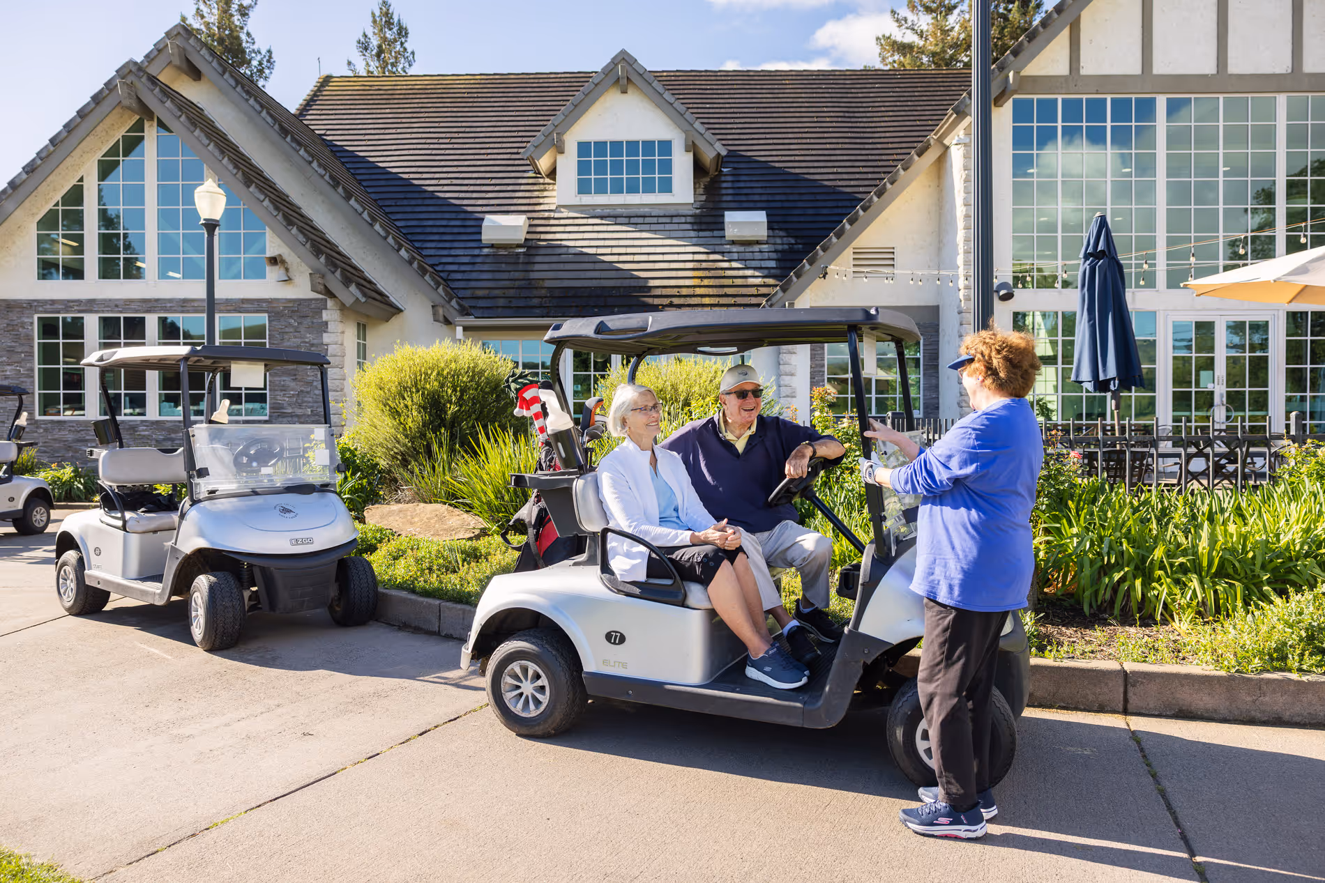 Two people sit in a golf cart talking to a woman standing nearby.