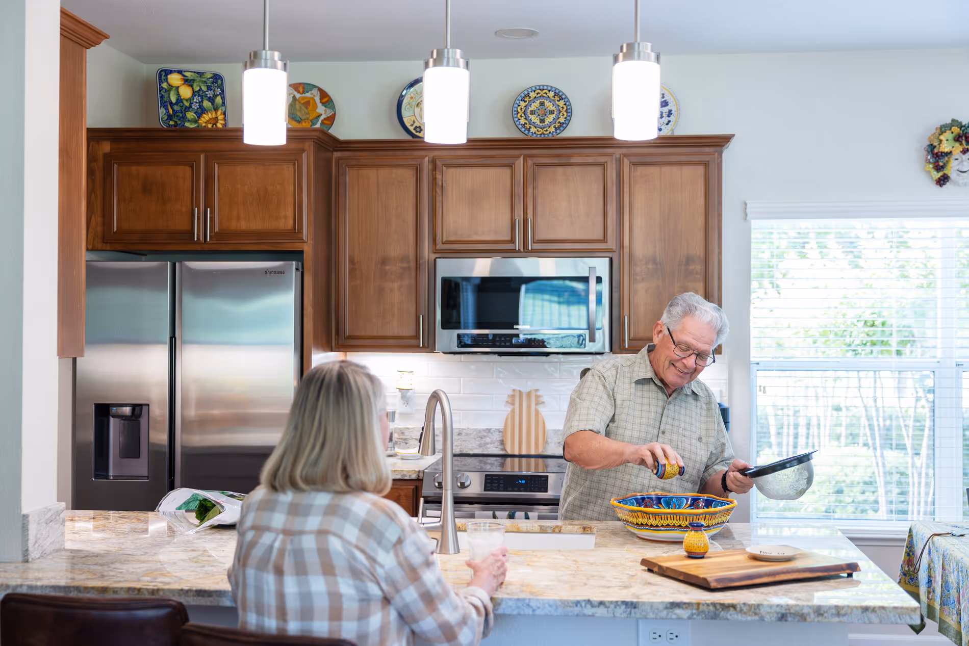 A man in a kitchen serves food into a bowl as a woman watches from the counter.