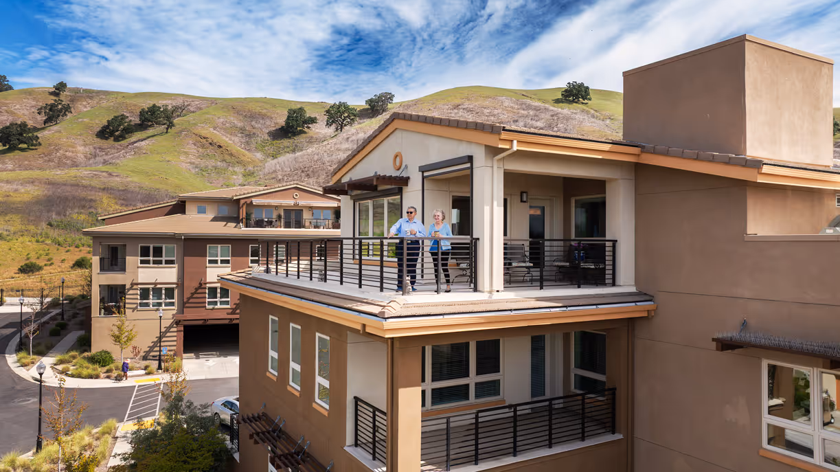 Two people stand on a large balcony of a modern building with hills in the background.