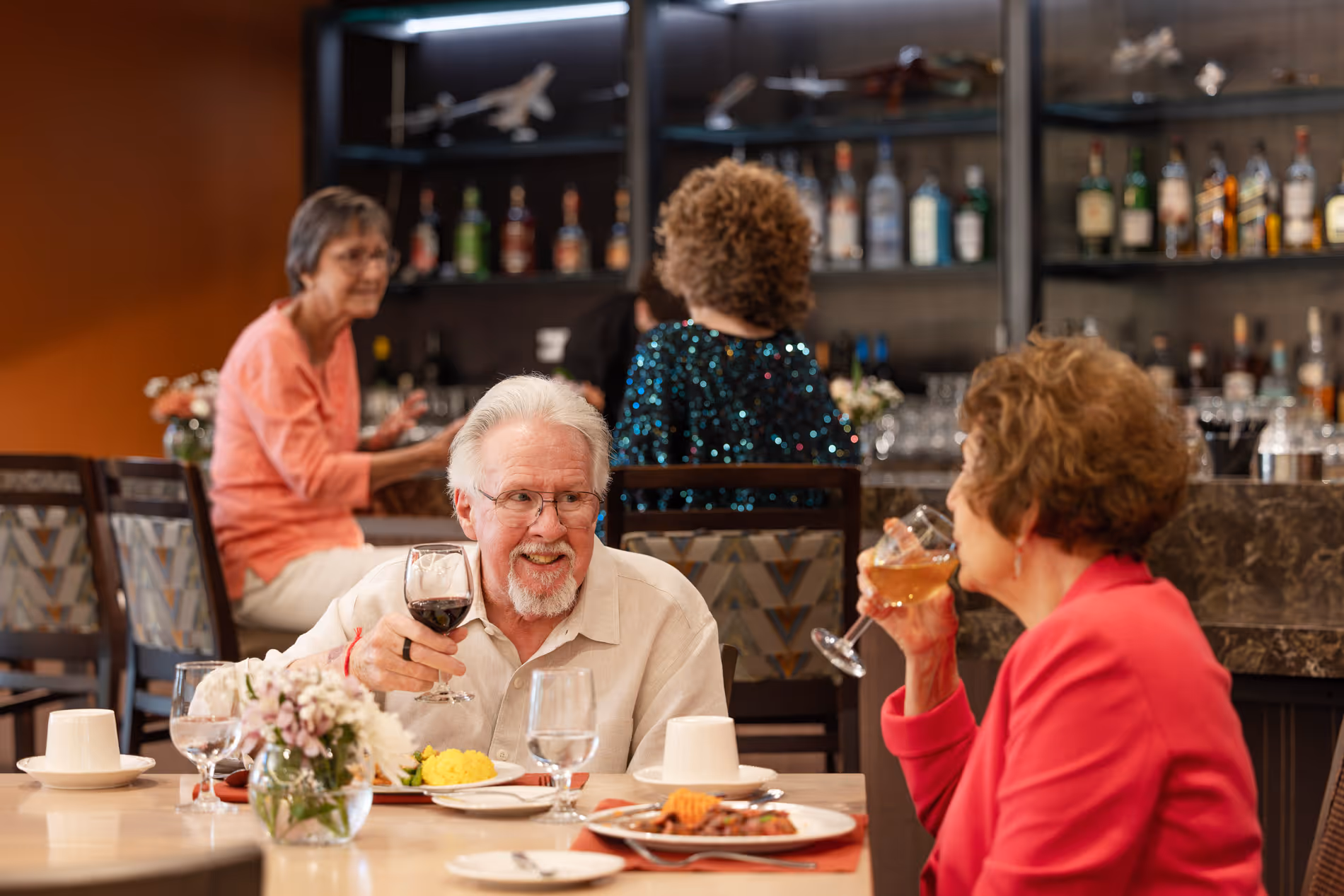 An older couple drinks wine and smiles at a restaurant table. Other patrons sit at the bar.