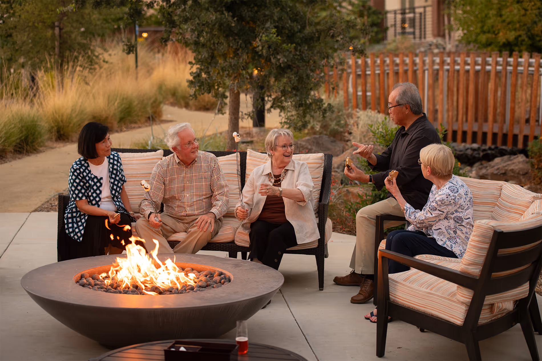 Five seniors sitting around fire pit roasting marshmallows and eating s'mores outdoors.