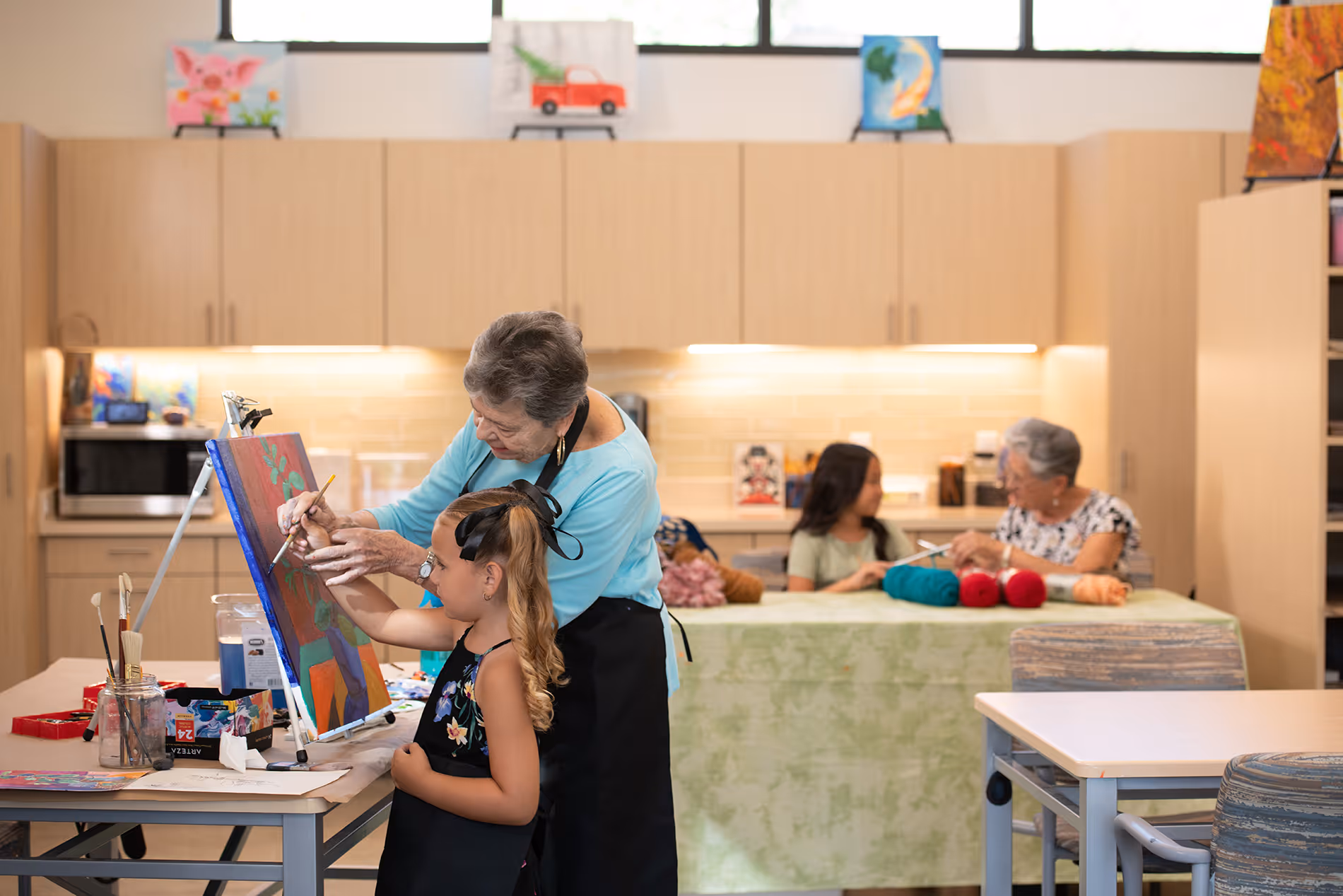 An older woman helps a young girl paint on a canvas in a classroom setting.