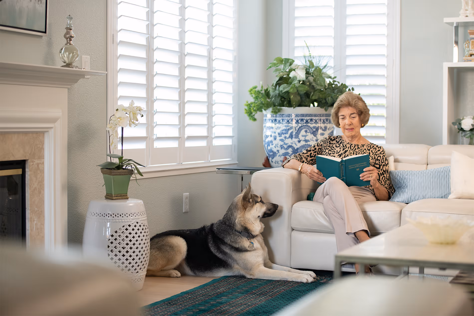 An older woman reads a book on a sofa while a large dog lies on the floor nearby.