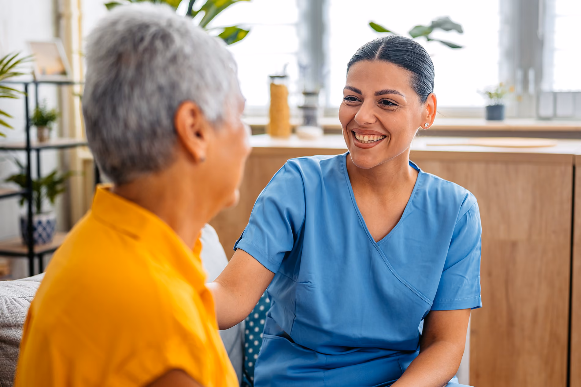 A healthcare worker in blue scrubs smiles and talks with an elderly woman wearing a yellow shirt.