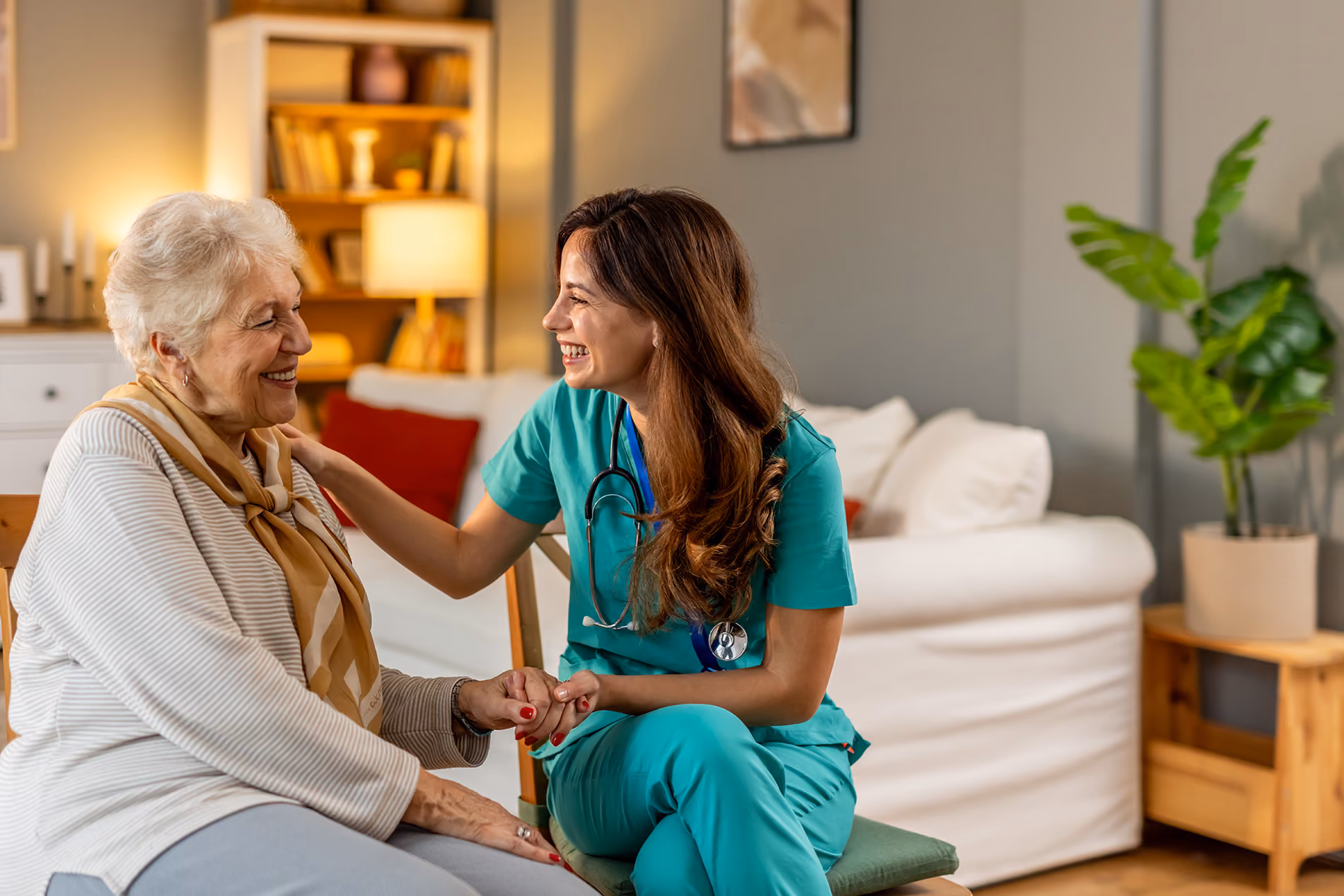 A nurse in scrubs smiles and talks with an elderly woman, holding her hand for support.