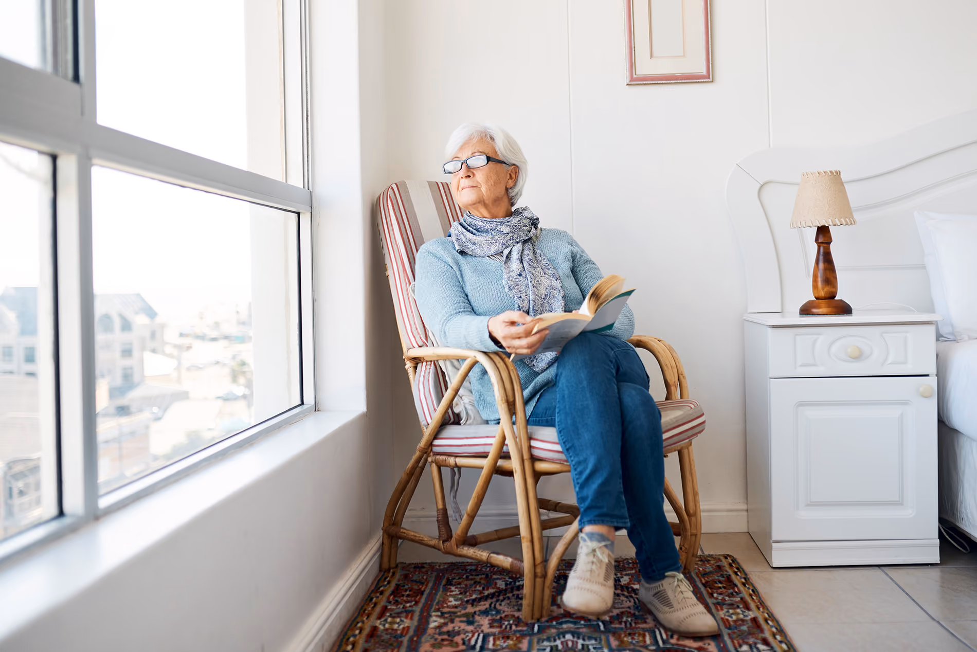 Elderly person sitting in a chair by a window, holding an open book, looking outside.