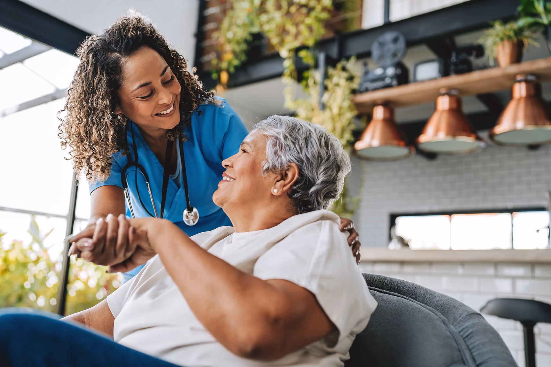 A nurse smiles while helping an elderly woman in a comfortable home setting.