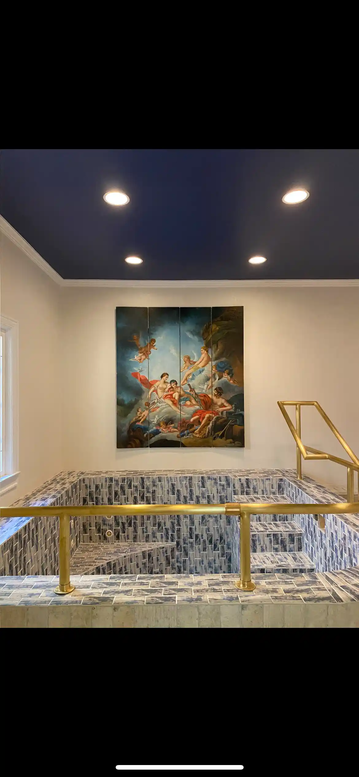Tiled indoor soaking pool or spa with blue ceiling, brass railings, and wall-mounted artwork above the water feature