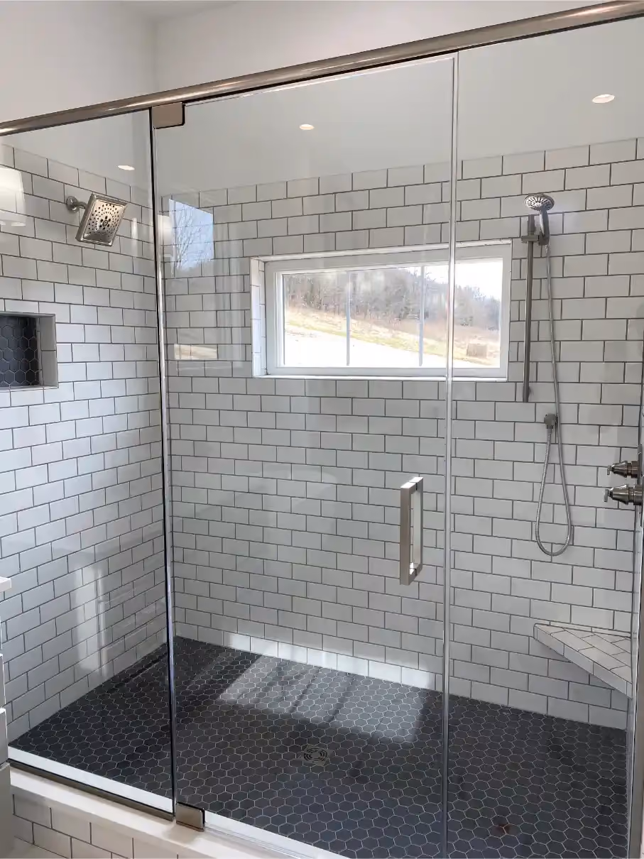 Glass-enclosed walk-in shower with white subway tile walls, gray hexagonal floor tile, dual shower heads, and a built-in bench