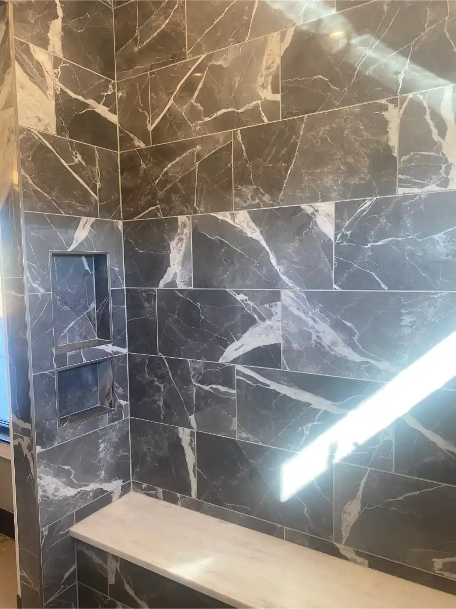 Close-up of a shower wall with large dark marble-look tiles featuring dramatic white veining, built-in niches, and a stone bench