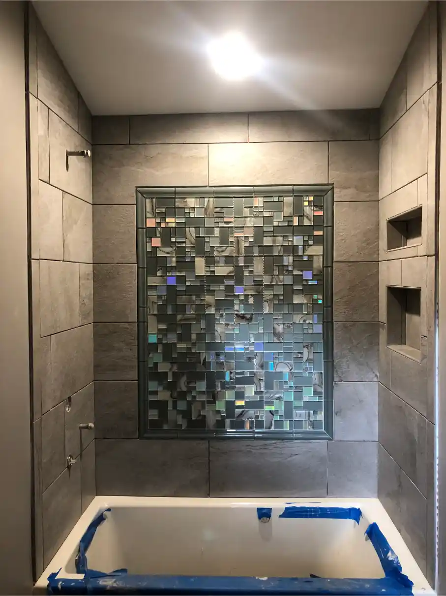 Bathtub alcove with large gray wall tiles, two recessed storage niches, and a decorative glass block window above the tub