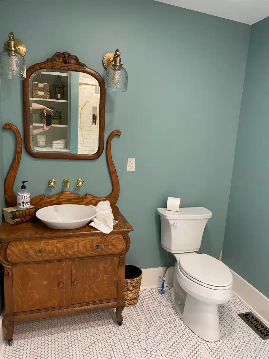Small bathroom with a vintage wood vanity, vessel sink, matching framed mirror, brass sconces, blue-green walls, and hex tile floor