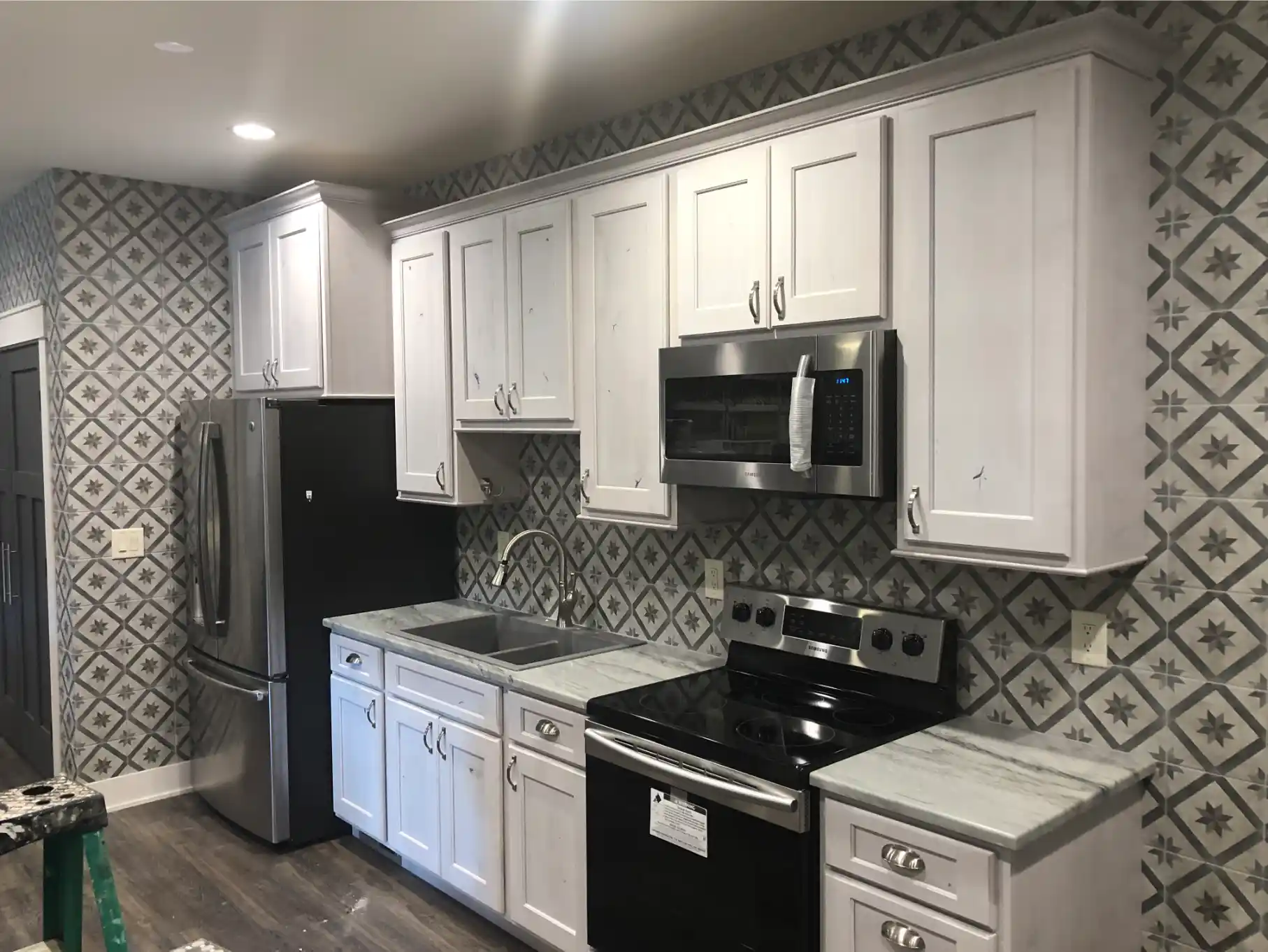 Compact kitchen with white cabinets, patterned full-wall tile backsplash, black range, stainless refrigerator, and gray countertops
