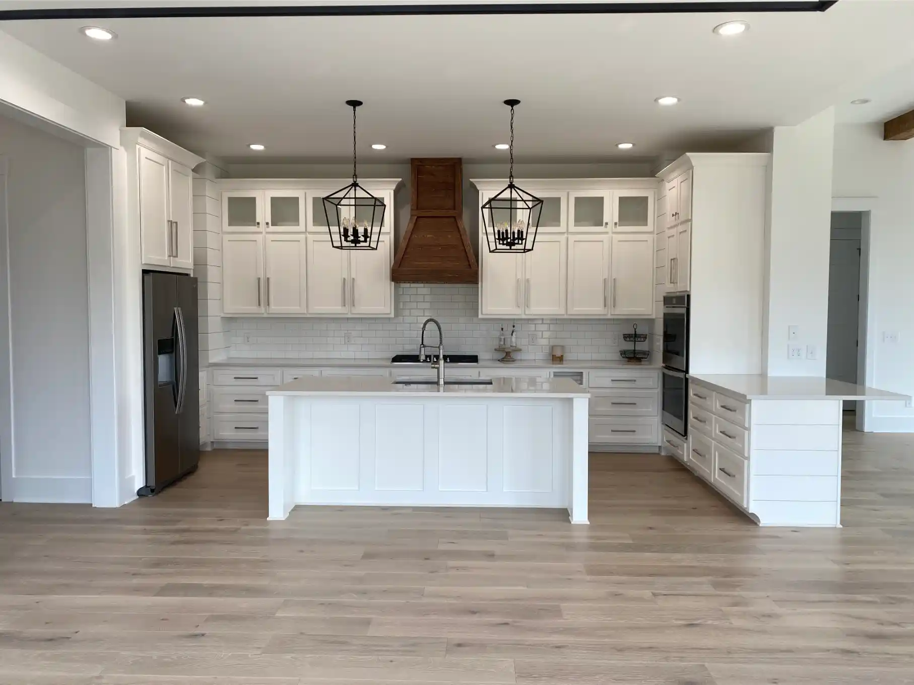 Wide view of a white open-concept kitchen with central island, wood range hood, black lantern pendants, and light wood flooring