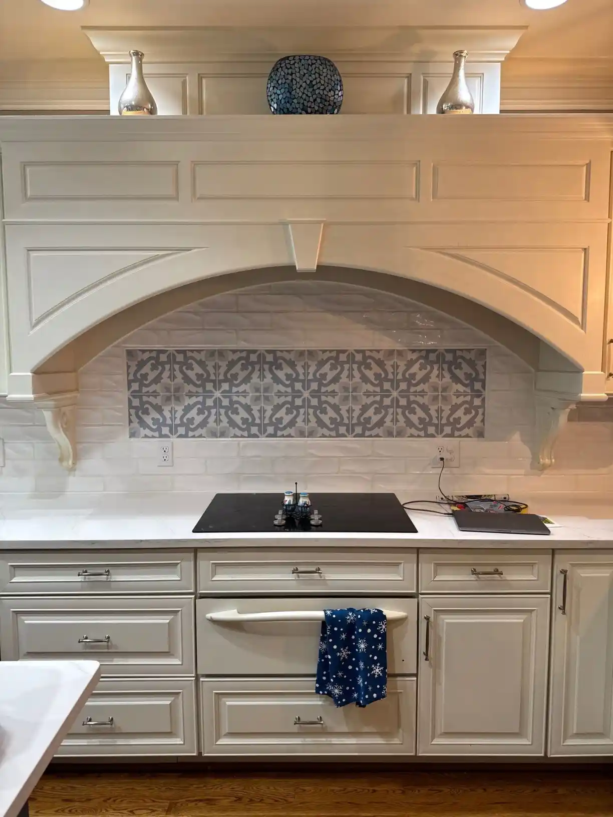 Decorative kitchen range alcove with white cabinetry, arched wood trim hood surround, patterned accent tile, and white subway tile backsplash