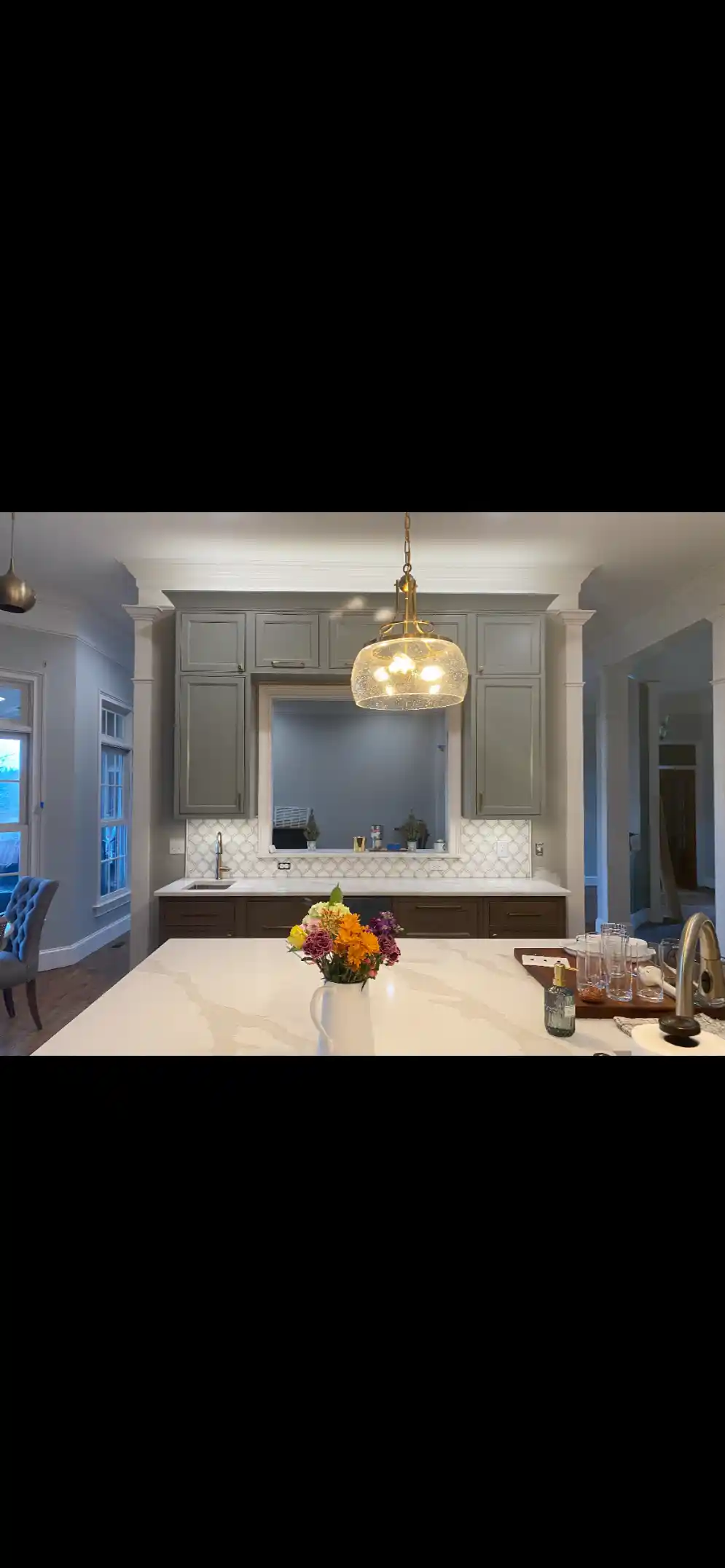 Kitchen with gray upper cabinets, white island countertop, gold pendant light, and a tiled backsplash viewed from across the island