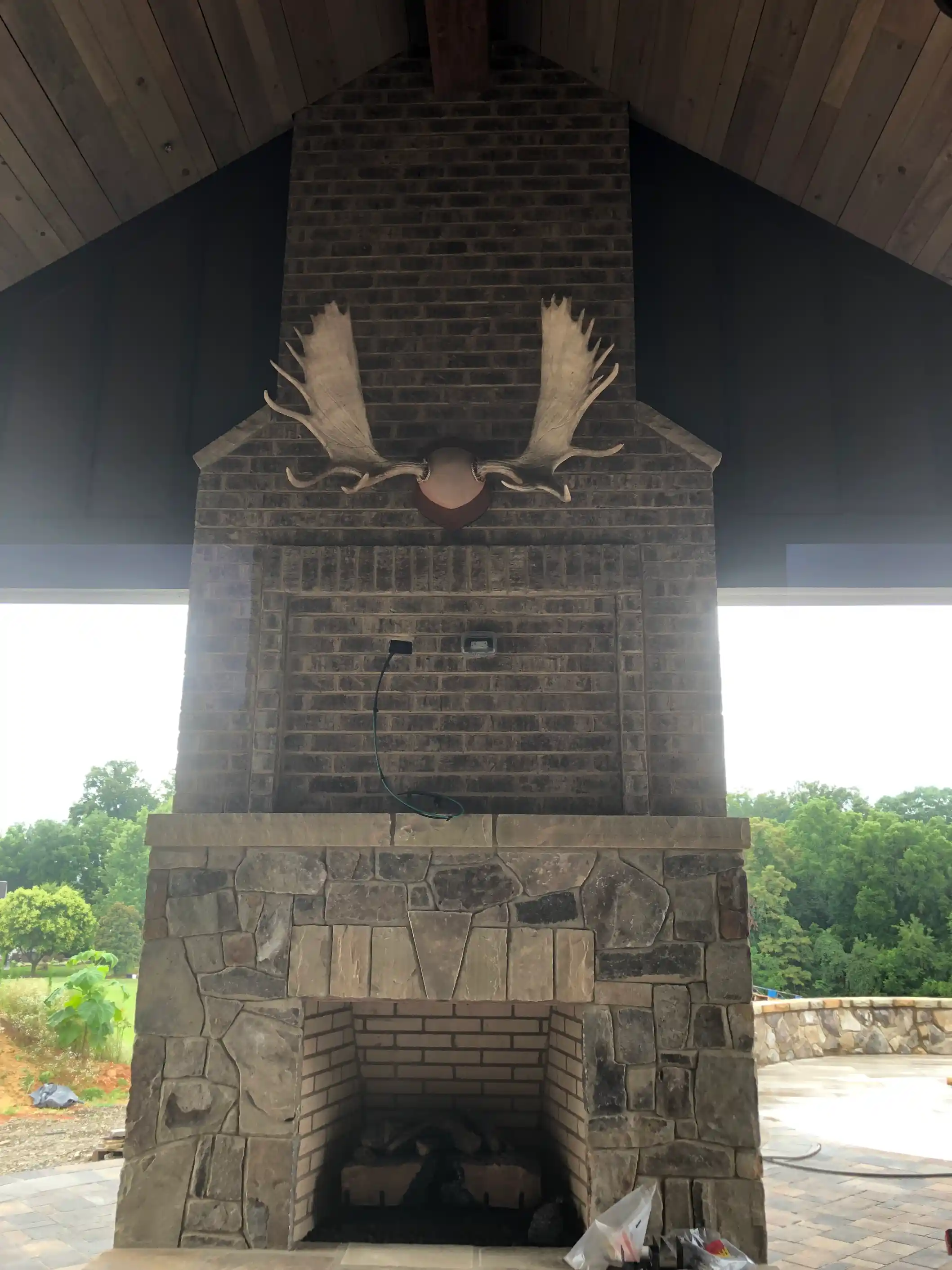 Tall outdoor stone fireplace with a brick chimney, vaulted wood ceiling, and mounted moose antlers in a covered patio area