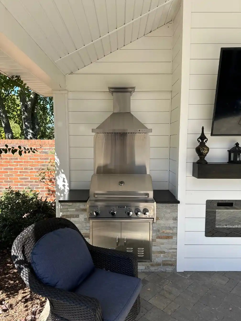 Covered outdoor kitchen with a built-in stainless grill, white shiplap walls, stone base, and adjacent wall-mounted TV