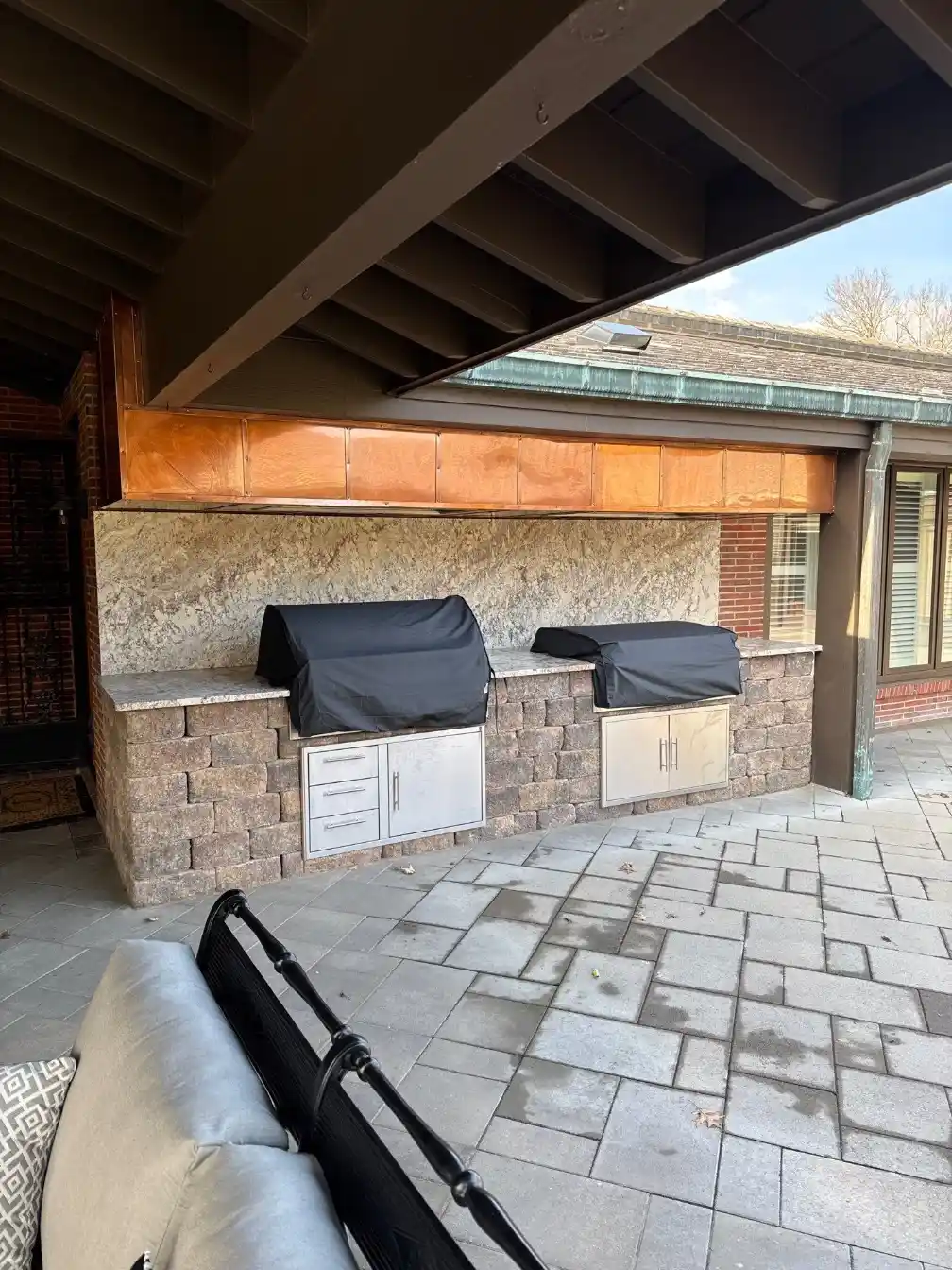 Covered patio outdoor kitchen with built-in grills, stone block base, granite countertop, copper hood surround, and paved flooring