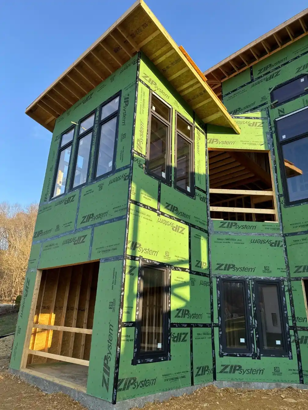 Exterior corner of a modern home under construction with ZIP System sheathing, black-framed windows, and exposed framing at the lower level