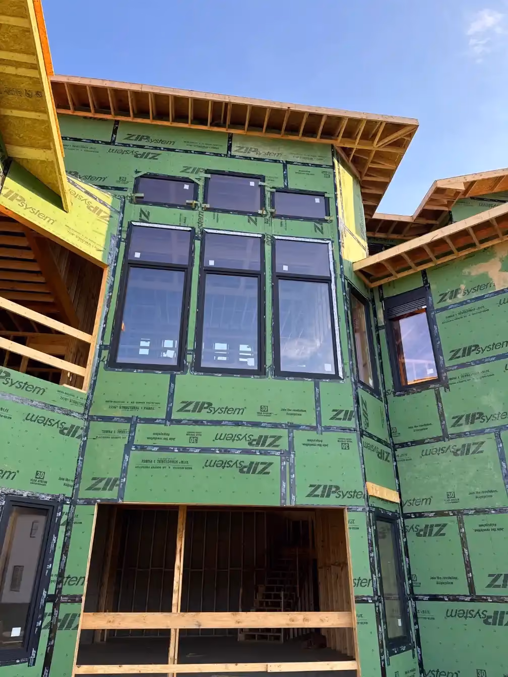 Two-story exterior wall under construction with ZIP System sheathing, a grid of black-framed windows, and an open lower-level framed entry