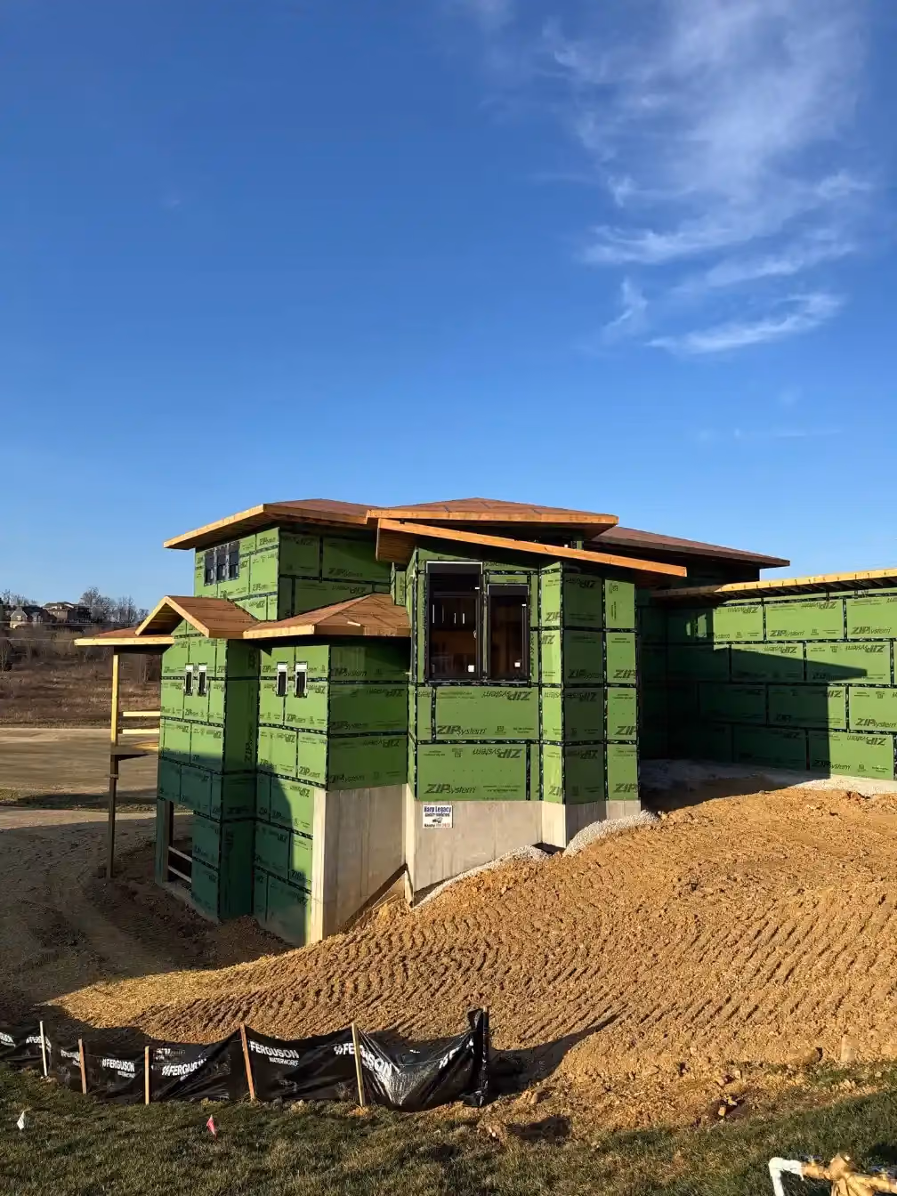 Wide exterior view of a custom home under construction with green ZIP System sheathing, multiple rooflines, and unfinished grading around the site